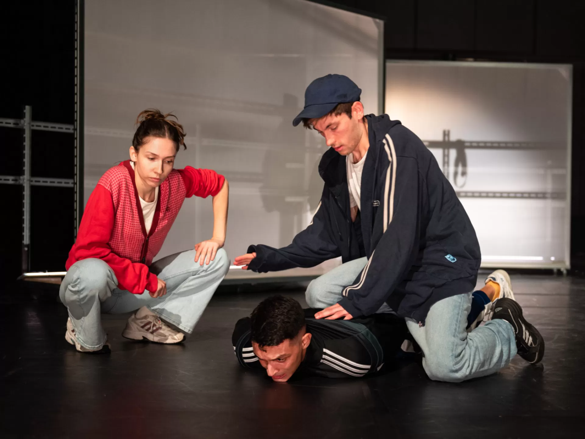 Auf dem Bild sind drei Personen in einem Bühnenraum zu sehen. Rechts zwei Männer, einer davon liegt bäuchlings am Boden, der andere kniet mit einem Knie auf ihm und drückt ihn nieder. Links daneben hockt eine Frau in Jeans, Turnschuhen und roter Jacke, die auf den am Boden liegenden Mann blickt.