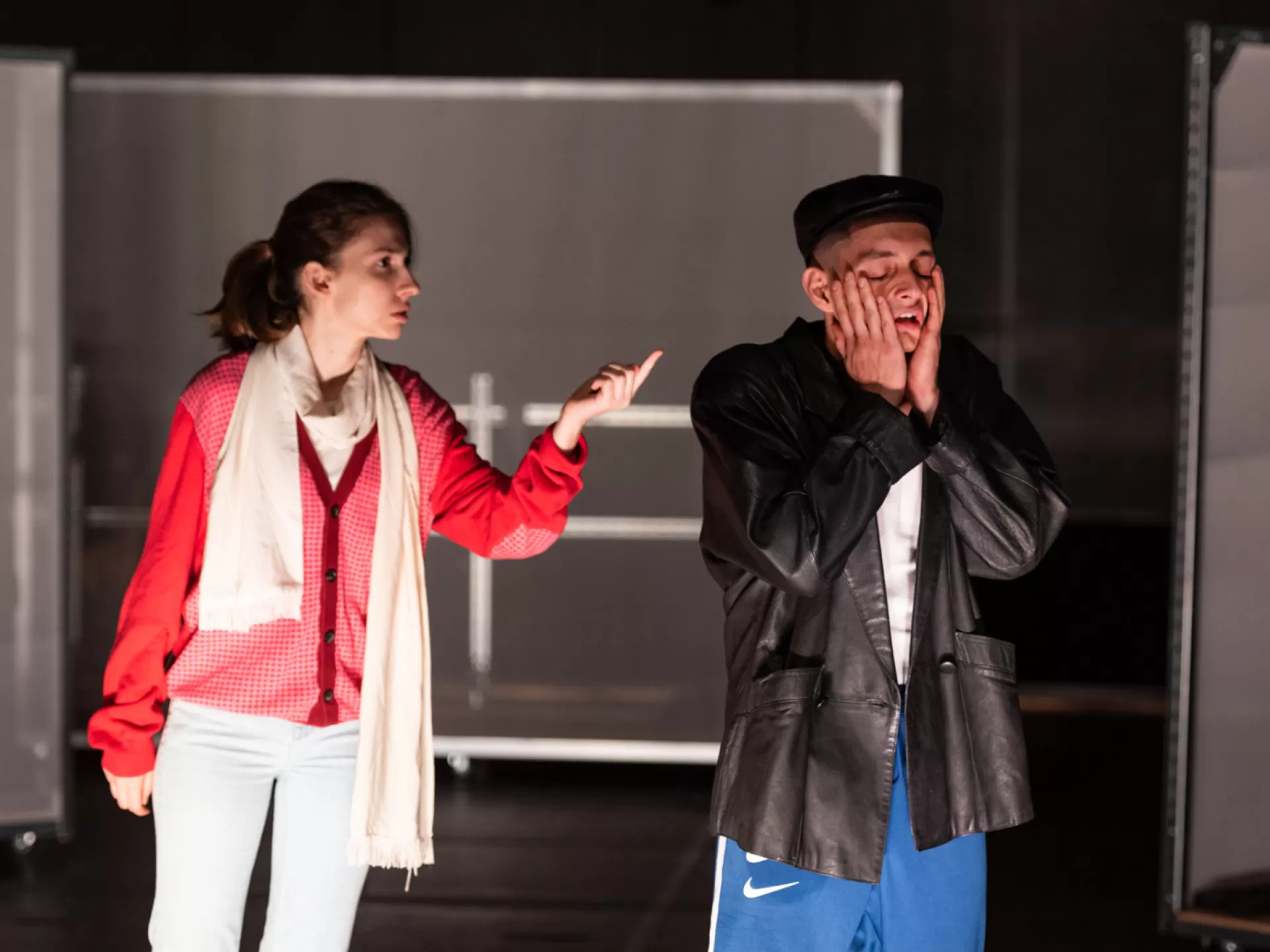 Zwei Personen stehen nebeneinander, sie sind zu sehen bis zum Knie. Links eine Frau mit hellem Schal, weißer Hose und roter Jacke. Sie hat die linke Hand gehoben und zeigt mit dem Zeigefinger auf einen Mann, der rechts neben ihr steht. Der Mann trägt eine schwarze Lederjacke und blaue Nike Sporthose, er hat die Hände seitlich an seine Wangen geschlagen und blickt verzweifelt.
