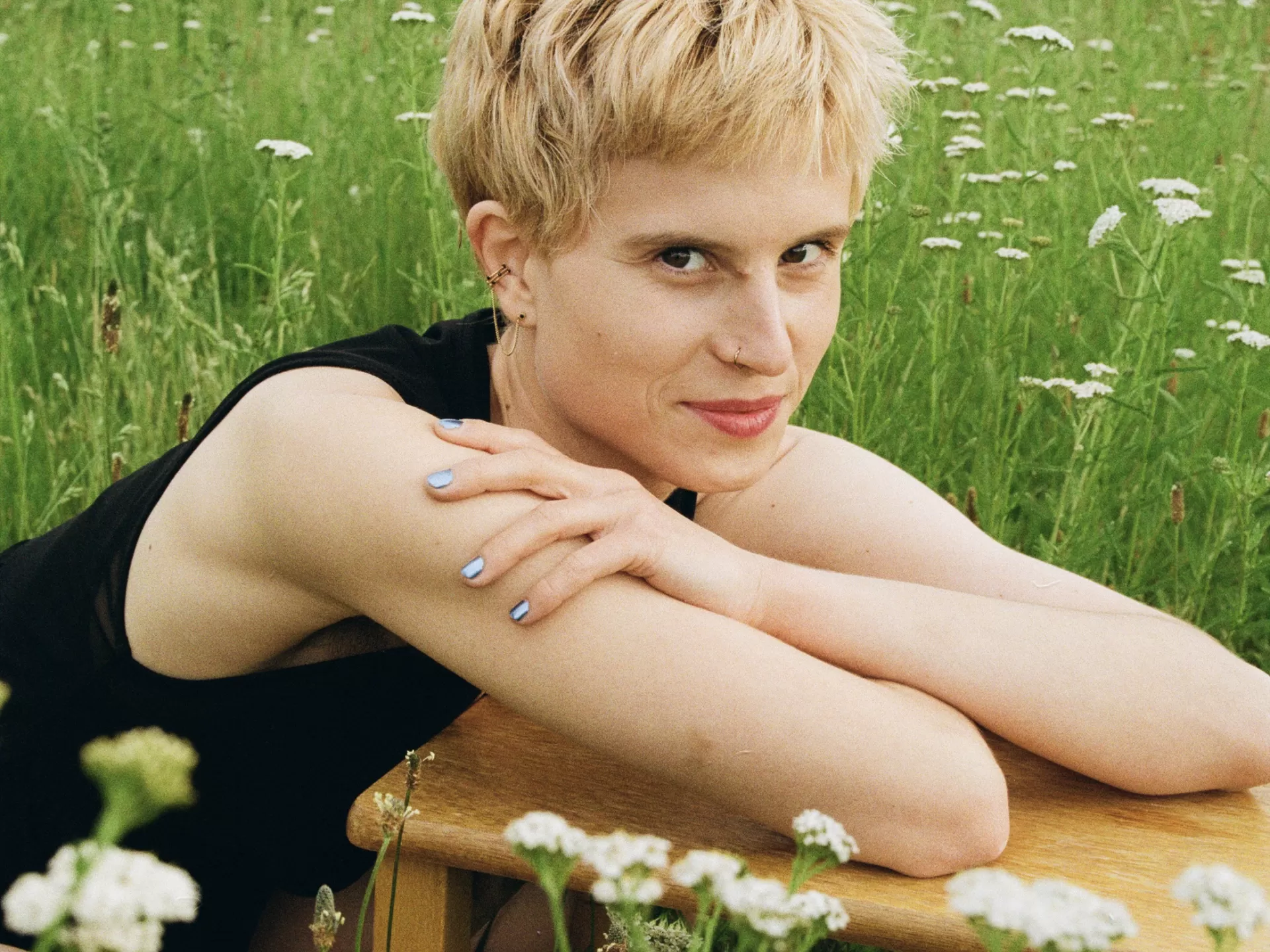Lisa lehnt sitzend mit den Händen auf einem Hocker. Sie ist in einer grünen Wiese. trägt ein schwarzes Kleid und blickt verschmitzt in die Kamera.