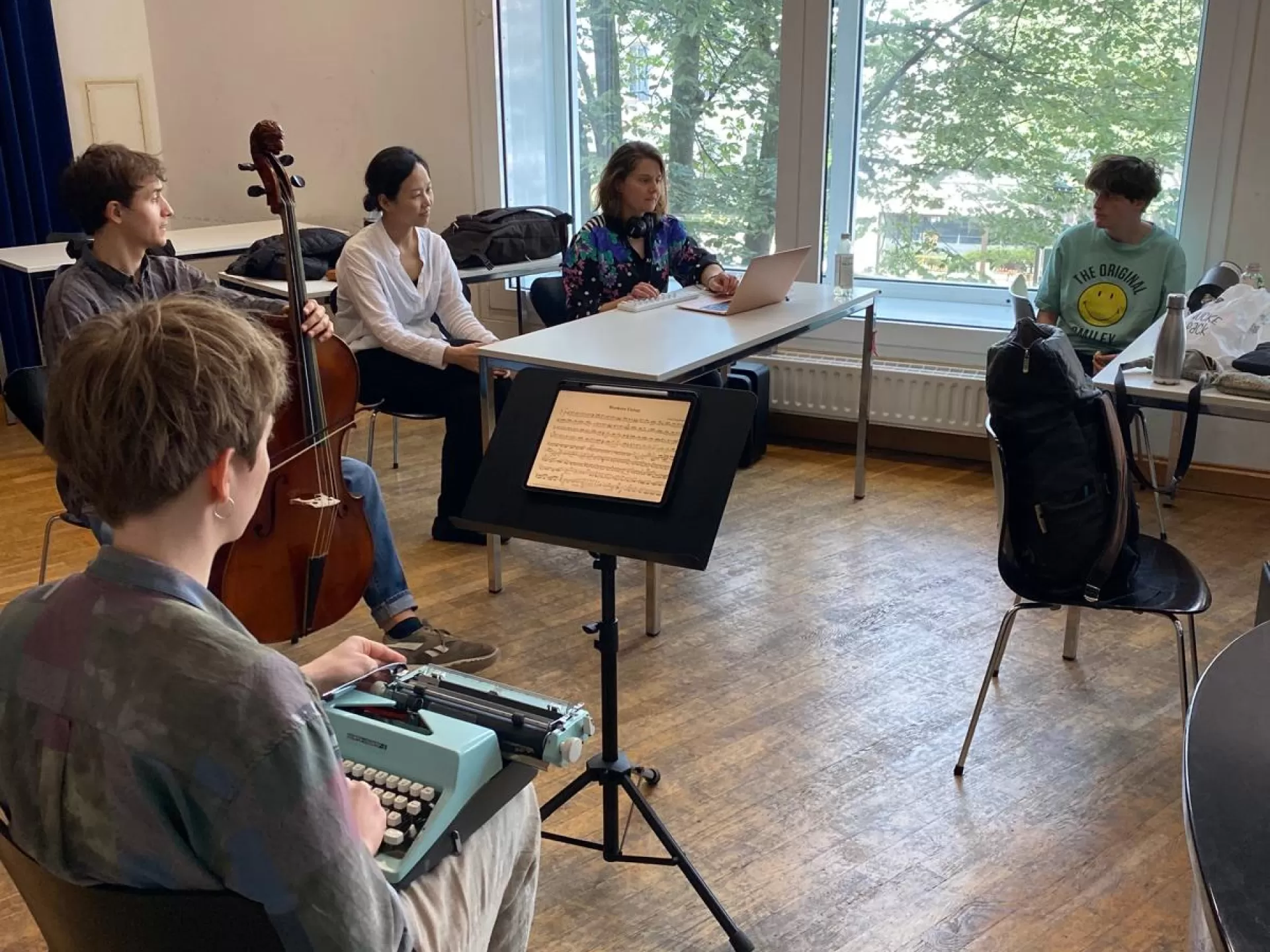 Fünf Studierende sitzen in einer Probe zusammen.  Eine Studentin mit kurzen braunen Haaren sitzt links mit dem Rücken zur Kamera im Bildvordergrund. Sie hat eine Schreibmaschine auf dem Schoß. Hinter ihr sitzt ein Student mit einem Cello. Die drei übrigen Studierenden sitzen an Tischen. Sie unterhalten sich.