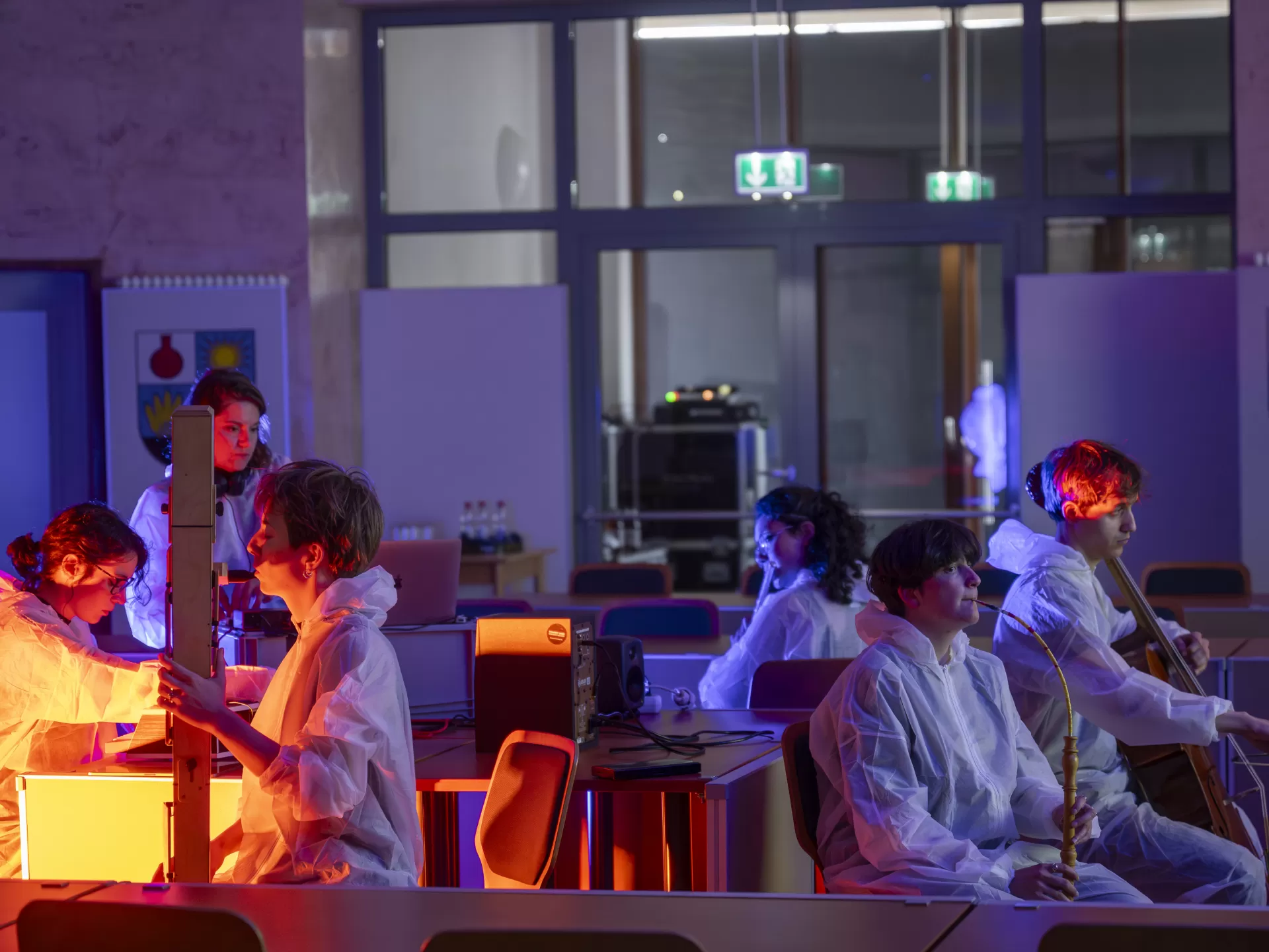 Eine Gruppe von Musiker*innen sitzt um einen Tisch im Rathaus herum. Alle tragen Einwegoveralls, wie sie die Spurensicherung an einem Tatort trägt. Sie spielen auf ihren Instrumenten und schauen in unterschiedliche Richtungen. Von links kommt orangenes Licht, der Rest des Bildes ist in bläuliches Licht getaucht.