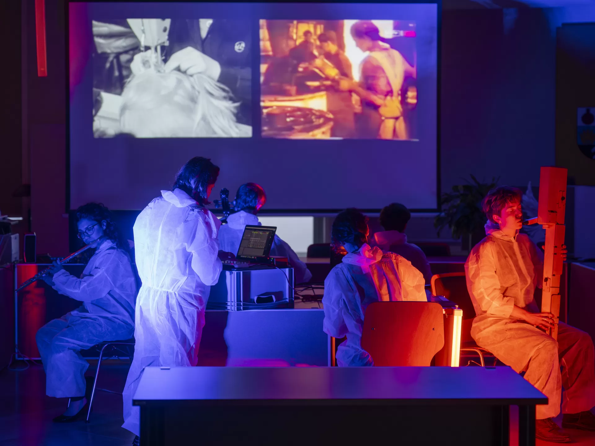 Eine Gruppe Musiker*innen sitzt mit verschiedenen Instrumenten um einen Tisch herum und musiziert. Alle tragen weiße Einweg-Overalls. Eine Studentin links im Bild steht an einem Laptop. Im Hintergrund sieht man historische Fotos, die an die Wand projiziert werden. Der Raum ist in blaues und rot-orangenes Licht getaucht.