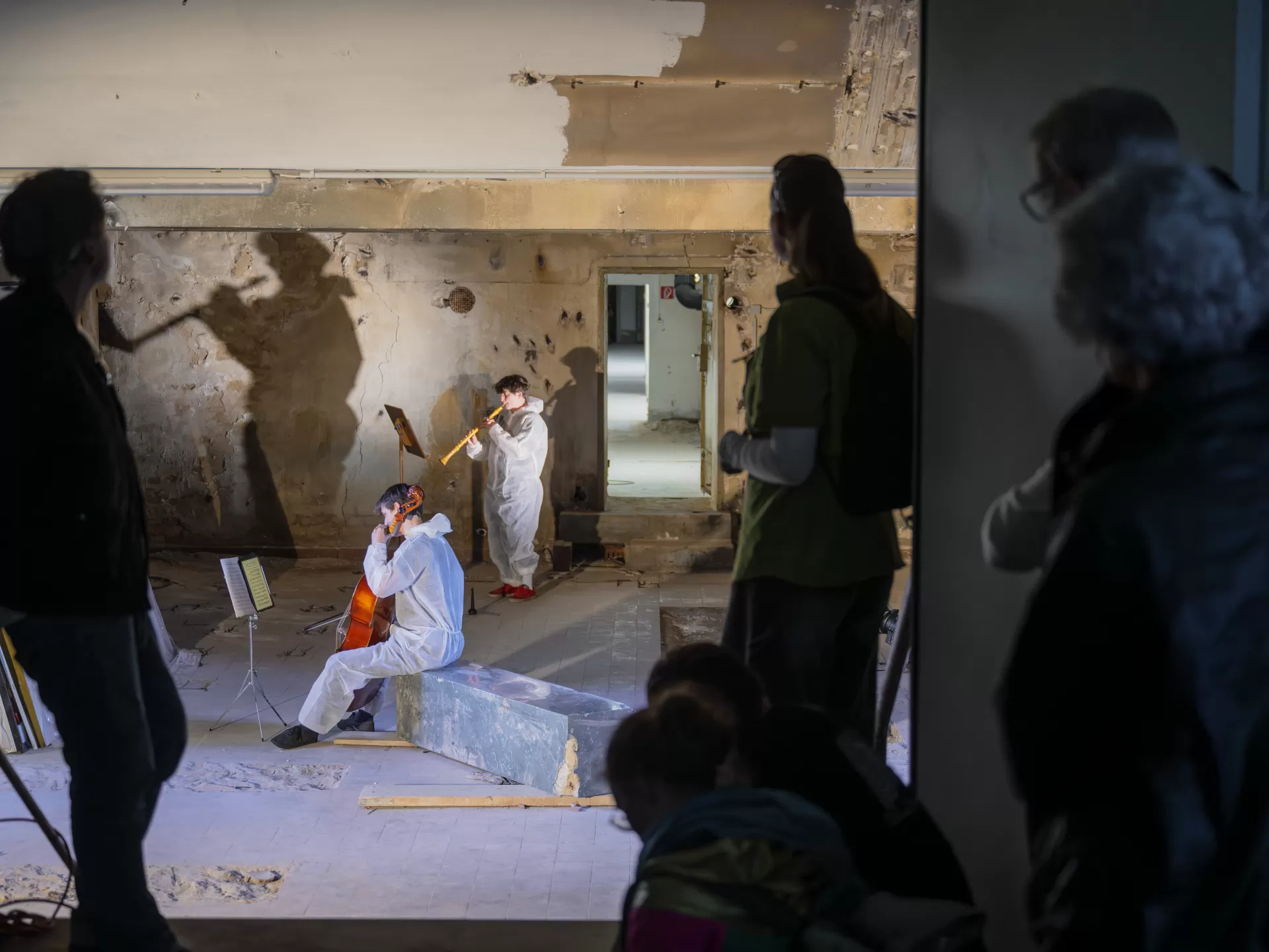 In einem Kellerraum musizieren zwei Studierende mit Cello und Oboe. Sie tragen weiße Einweg-Overalls. Durch das Licht werden ihre Schatten groß an die Wand geworfen. Im Vordergrund des Bilds sind einige Zuschauer*innen zu sehen, die durch eine Türöffnung in den Raum schauen.