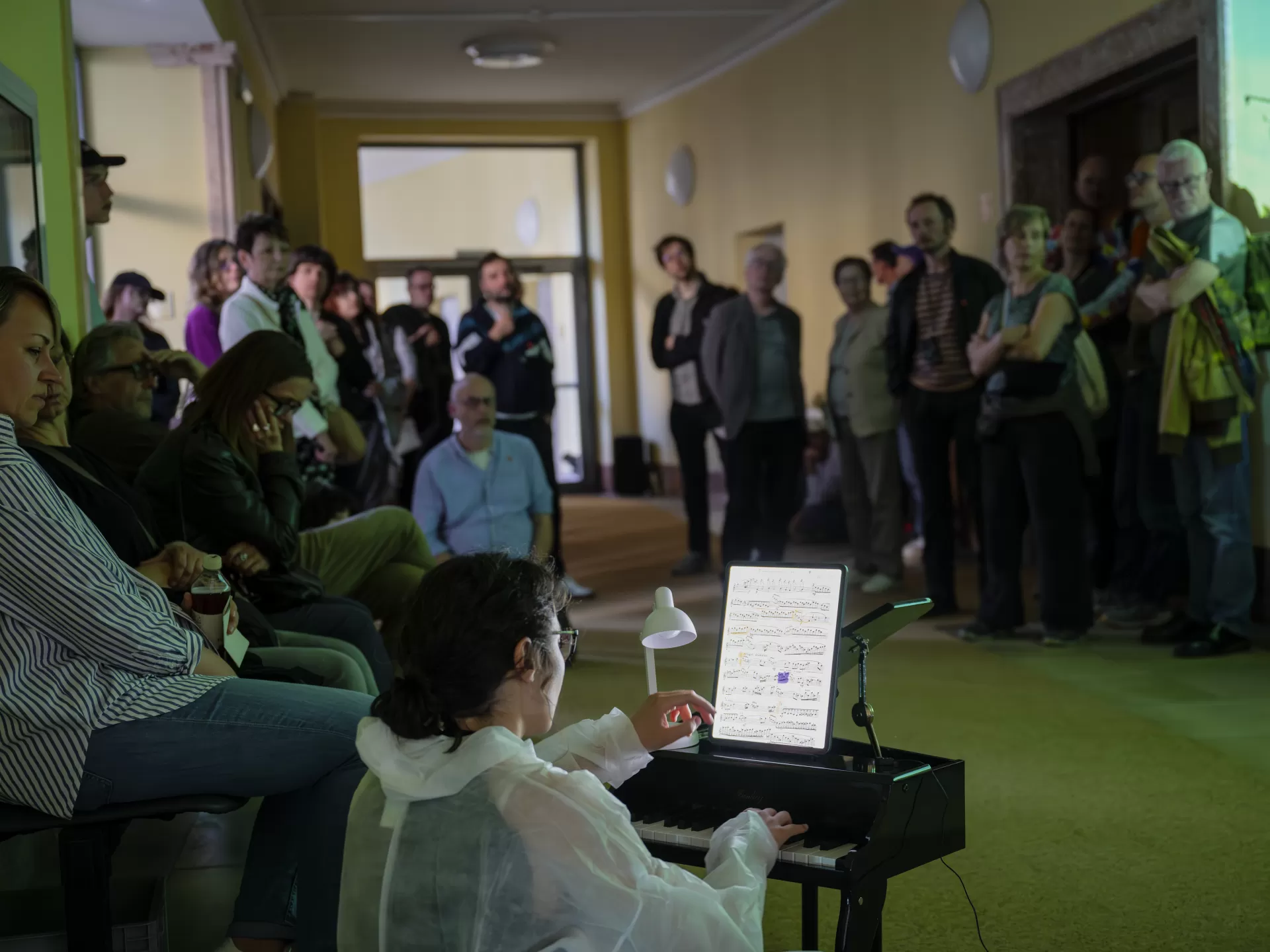 In einem Gang schaut eine Gruppe Zuschauer*innen, teilweise stehend, teilweise sitzend, auf eine Studentin unten links im Bild. Sie spielt ein winziges Klavier und trägt einen weißen Einweg-Overall. Die Noten hat sie auf einem Tablet. Rechts im Bild sieht man noch einen Teil einer Projektion an der Wand.