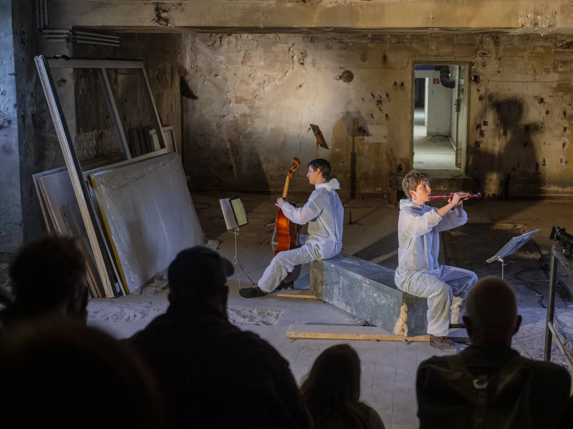 Ein Cellist und eine Blockflötistin sitzen auf einer Art rechteckigem Stein in einem baufällig aussehenden Keller und spielen. Im Anschnitt sind Zuschauer*innen zu sehen.