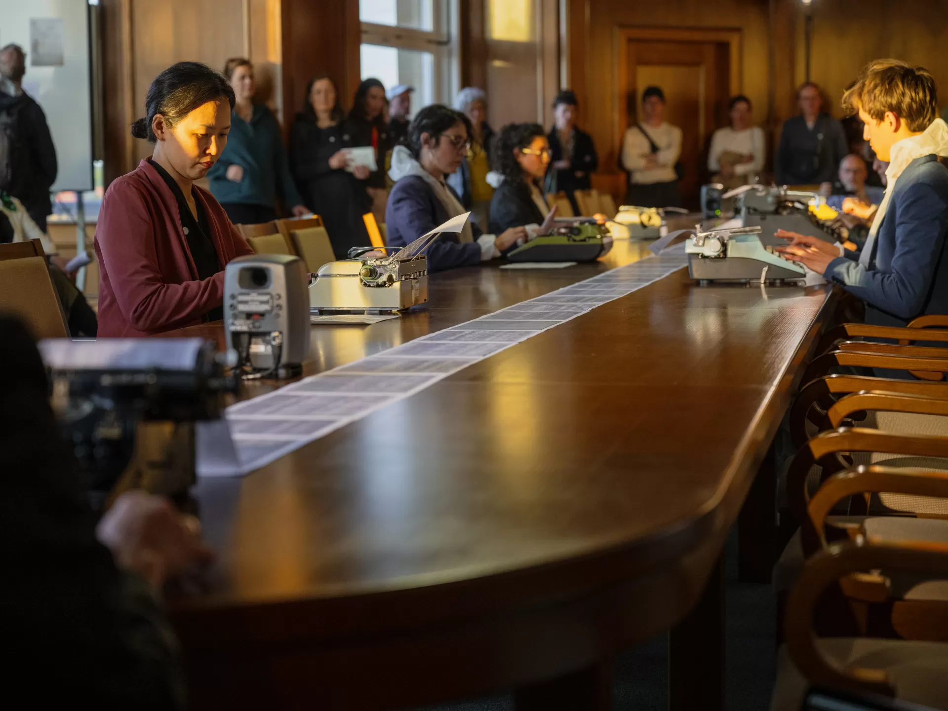 In einem Raum mit Holzvertäfelung sitzen die Studierenden an einem langen Holztisch verteilt und tippen auf Schreibmaschinen. Sie haben nun Alltagskleidung über ihre Overalls gezogen. Das Publikum steht am Rand und schaut zu.