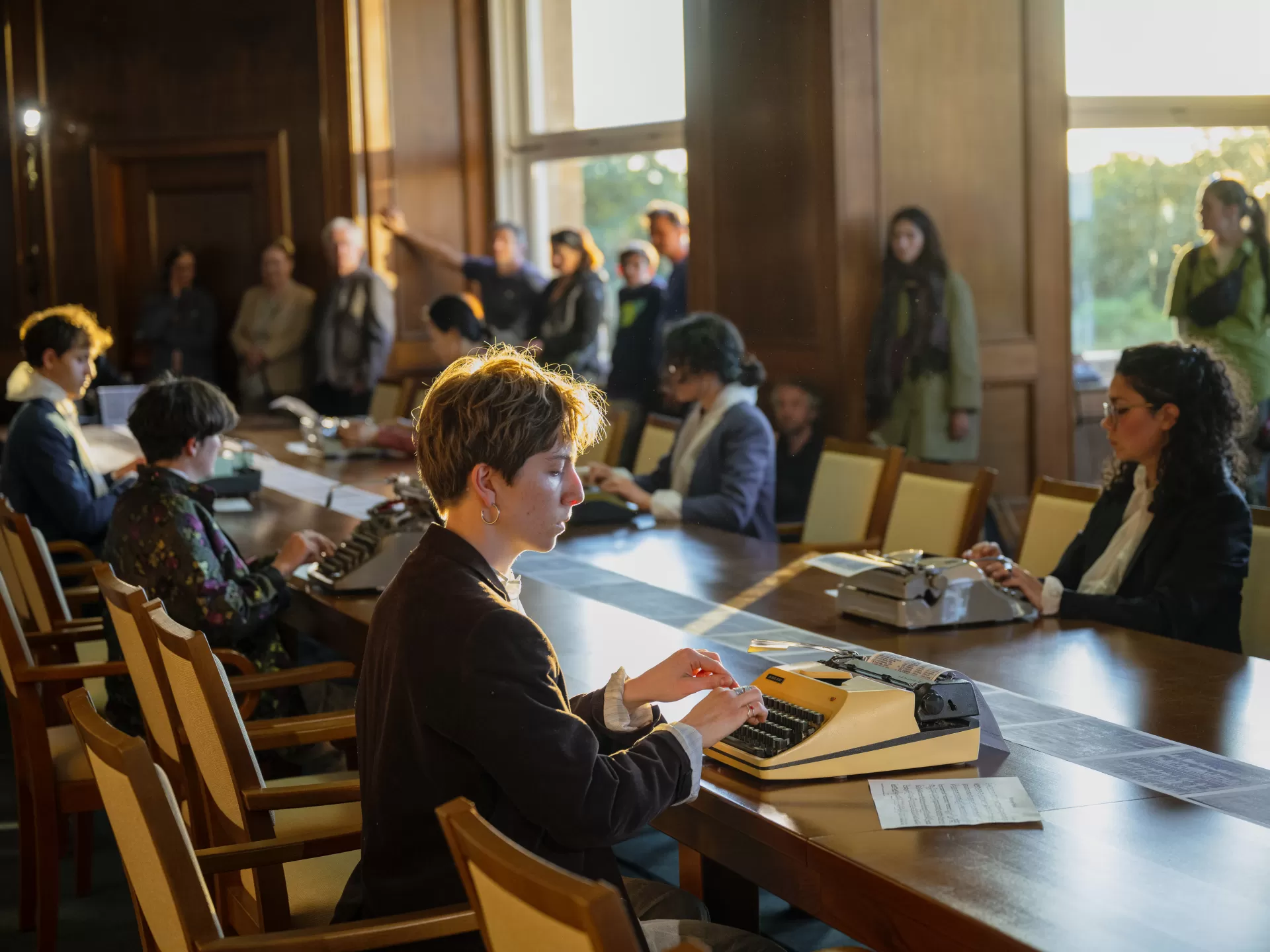 In einem Raum mit Holzvertäfelung sitzen die Studierenden an einem langen Holztisch verteilt und tippen auf Schreibmaschinen. Sie haben nun Alltagskleidung über ihre Overalls gezogen. Im Hintergrund sind große Fenster zu sehen. Das Publikum steht am Rand und schaut zu. Eine Studentin mit kurzen braunen Haaren und einem schwarzen Blazer ist hier im Vordergrund.