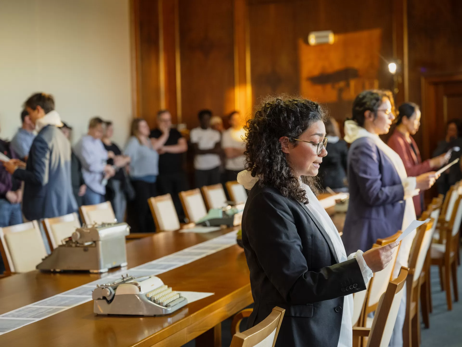 Die Studierenden stehen mit dem Rücken zu einem langen Holztisch und lesen etwas vor. Das Publikum steht am Rand und schaut zu.