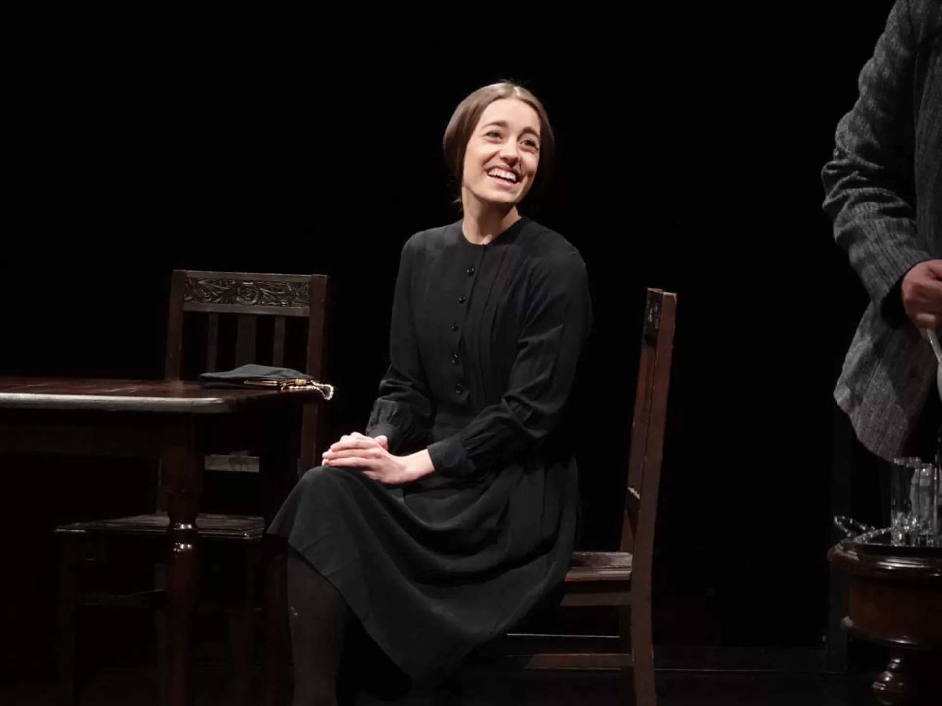 Eine Frau in dunkler Kleidung sitzt lächelnd auf einem Stuhl.