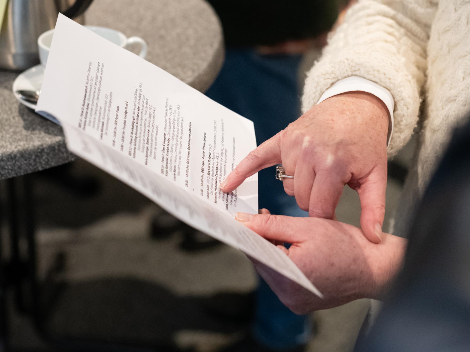Zwei Personen betrachten einen aufgeschlagenen Programmzettel, sichtbar sind dabei nur Hände: Die linke Hand der ersten Person hält das Blatt, die andere Person deutet mit dem kleinen Finger der rechten Hand auf einen Programmpunkt. Im Hintergrund sind andere Personen und Stehtische erkennbar, auf einem der Tische steht eine Kaffeekanne.