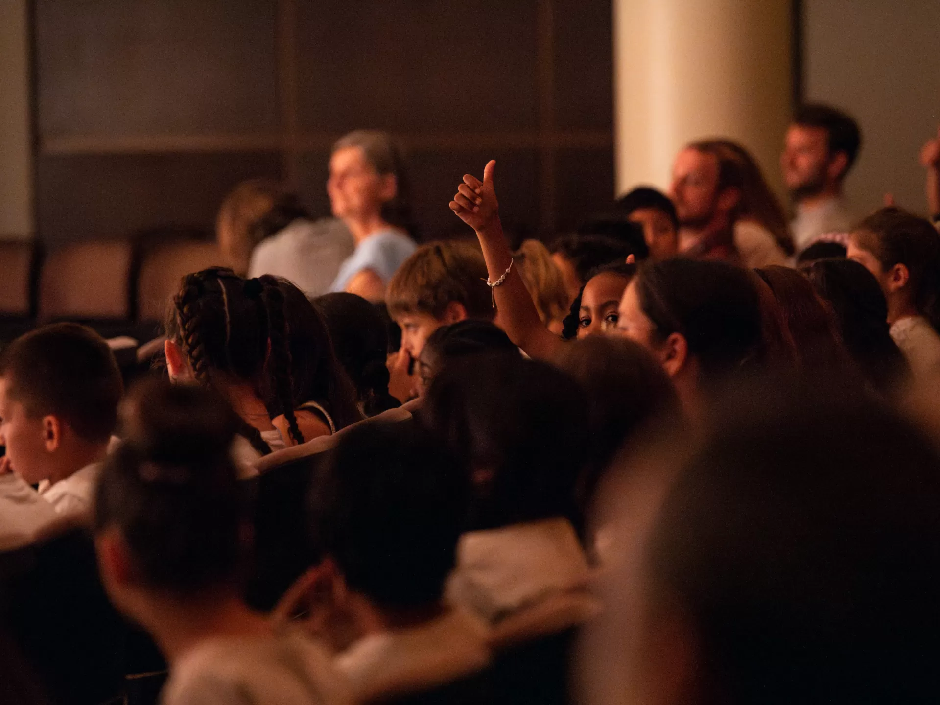 Das Publikum von schräg hinten fotografiert. Man sieht mehrere Reihen mit Kindern. Ein Mädchen hält ihren Daumen hoch und schaut nach links.