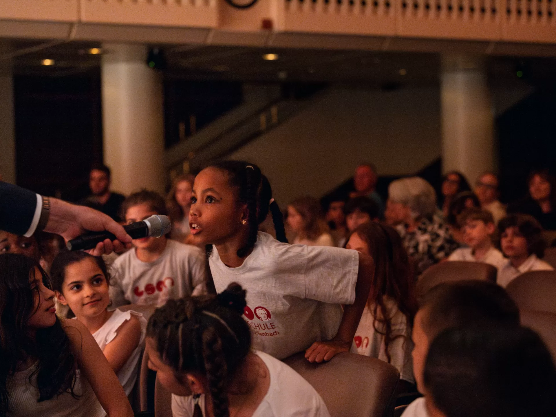 Ein Mädchen im Publikum steht hier im Fokus. Eine Hand aus dem Off hält ihr ein Mikrofon hin. Das Mädchen stützt sich auf den Stuhllehnen vor sich ab und lehnt sich nach vorne, um in das Mikrofon zu sprechen.