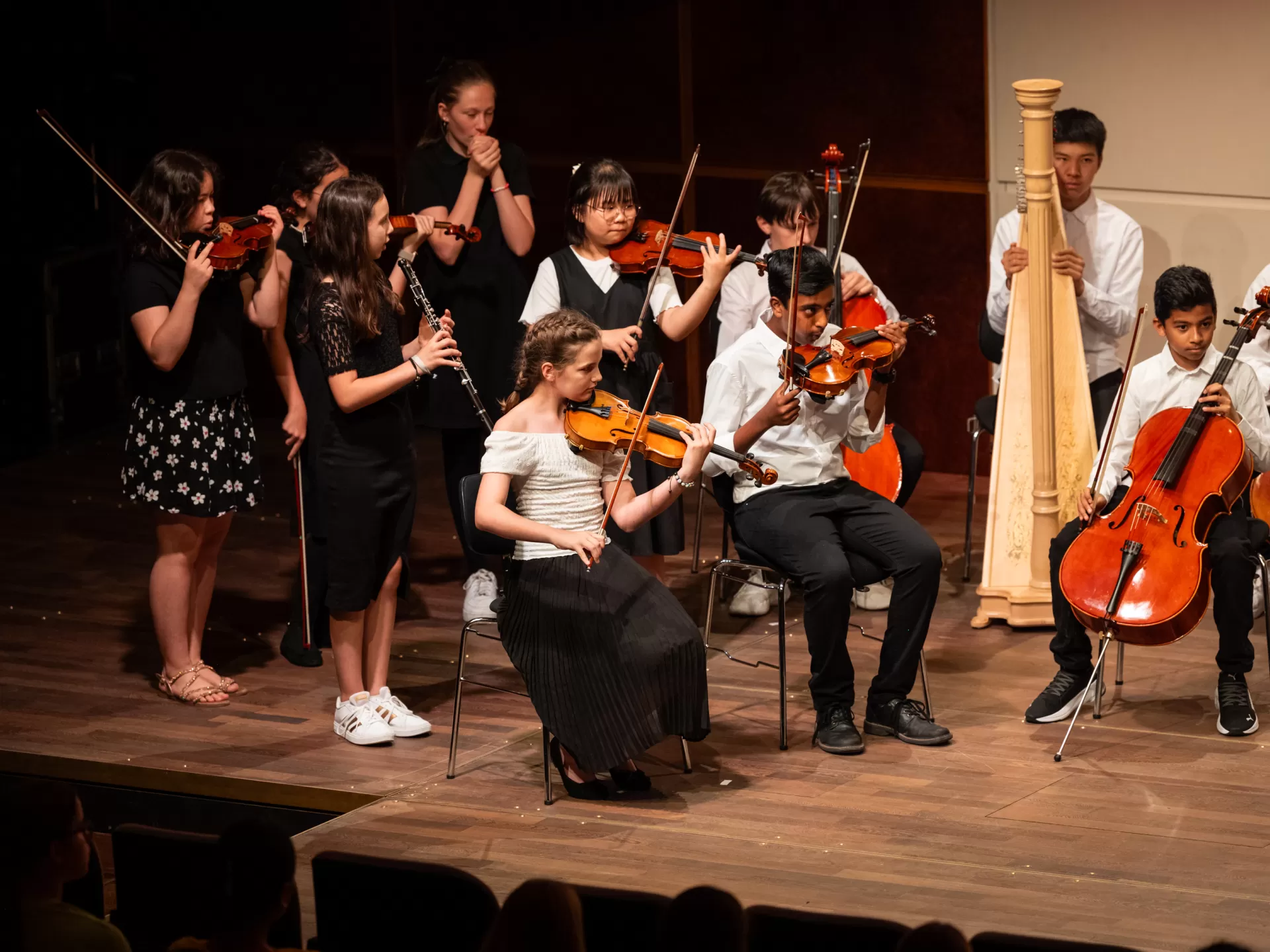 Eine Gruppe Kinder in Orchesterformation auf der Bühne. Die meisten haben Streichinstrumente. Sie tragen feine schwarz-weiße Kleidung und haben konzentrierte Gesichter. Manche von ihnen spielen gerade, die anderen scheinen auf ihren Einsatz zu warten.