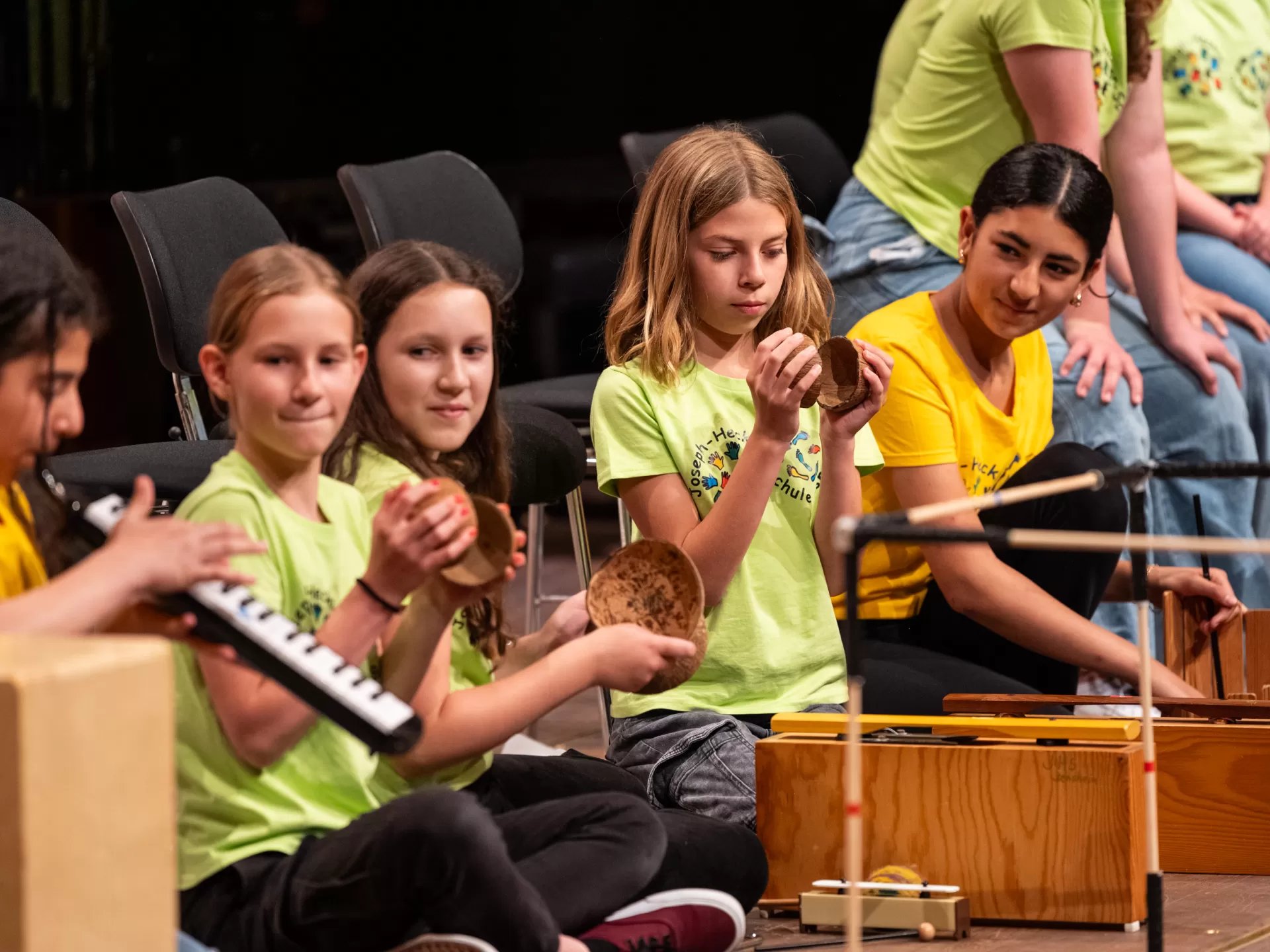 Eine Reihe von Schülerinnen kniet auf dem Boden mit verschiedenen Instrumenten. Sie tragen hellgrüne und gelbe T-Shirts. Drei der fünf Mädchen blicken nach links zu einem Mädchen, das gerade Melodica spielt. Der Rest hält Kokosnusshälften und andere Perkussionsinstrumente.