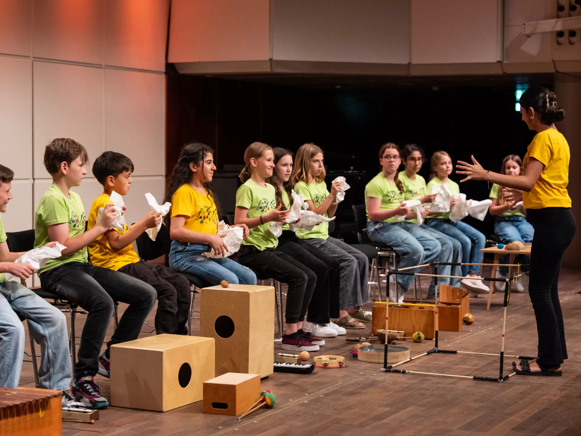 Kinder in hellgrünen und gelben T-Shirts sitzen in einer Reihe auf Stühlen und halten zerknülltes Papier in der Hand. Vor ihnen steht eine Dirigentin in einem gelben Shirt. Auf der Bühne liegen außerdem verschiedene Percussion-Instrumente.