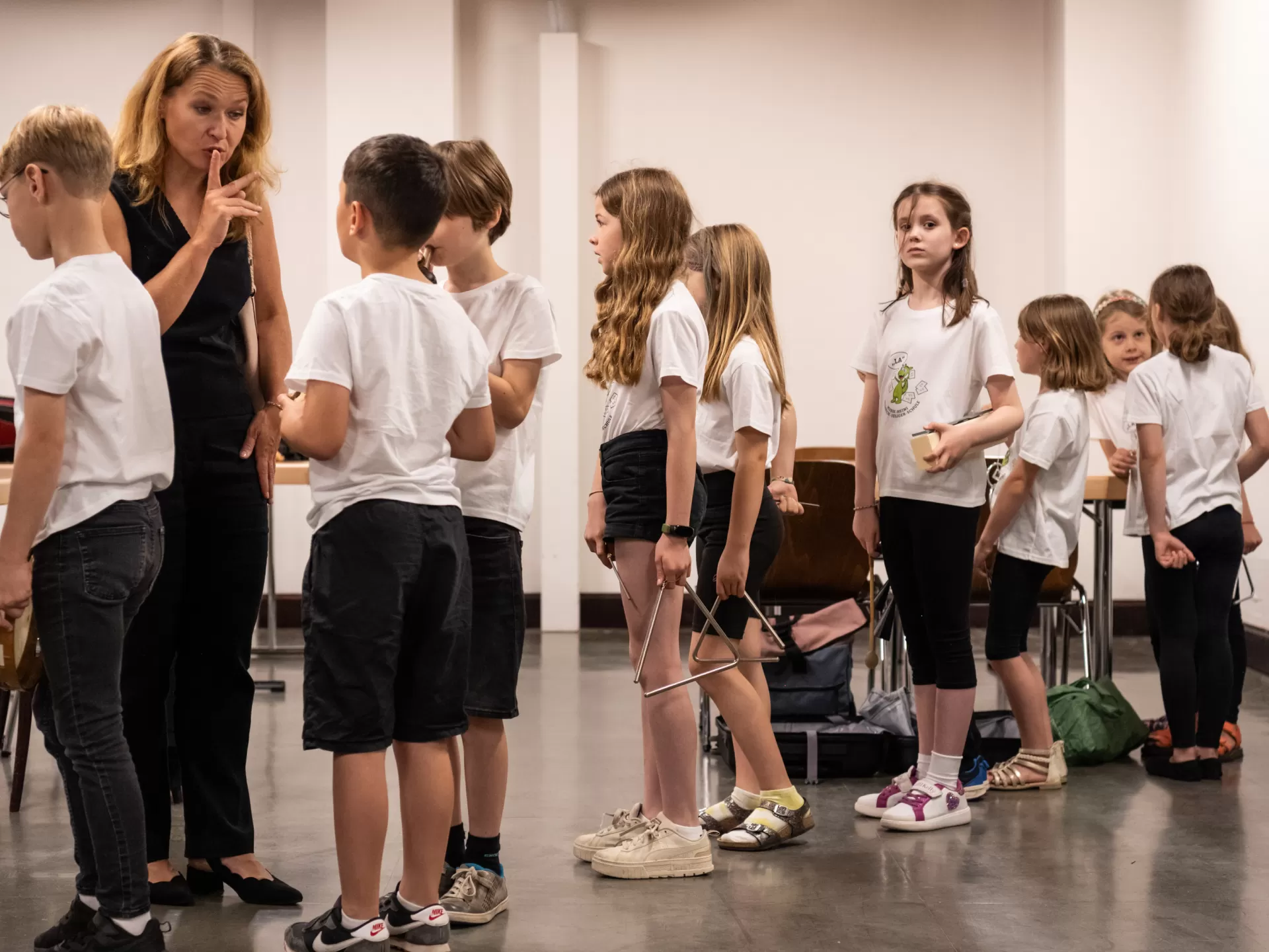 Hinter den Kulissen hat sich eine Grundschulklasse in weißen Shirts für den Auftritt bereit gemacht und steht in einer Schlange. Die blonde Lehrerin (links im Bild) legt ihren Finger auf die Lippen und bedeutet damit einem Schüler, ruhig zu sein.