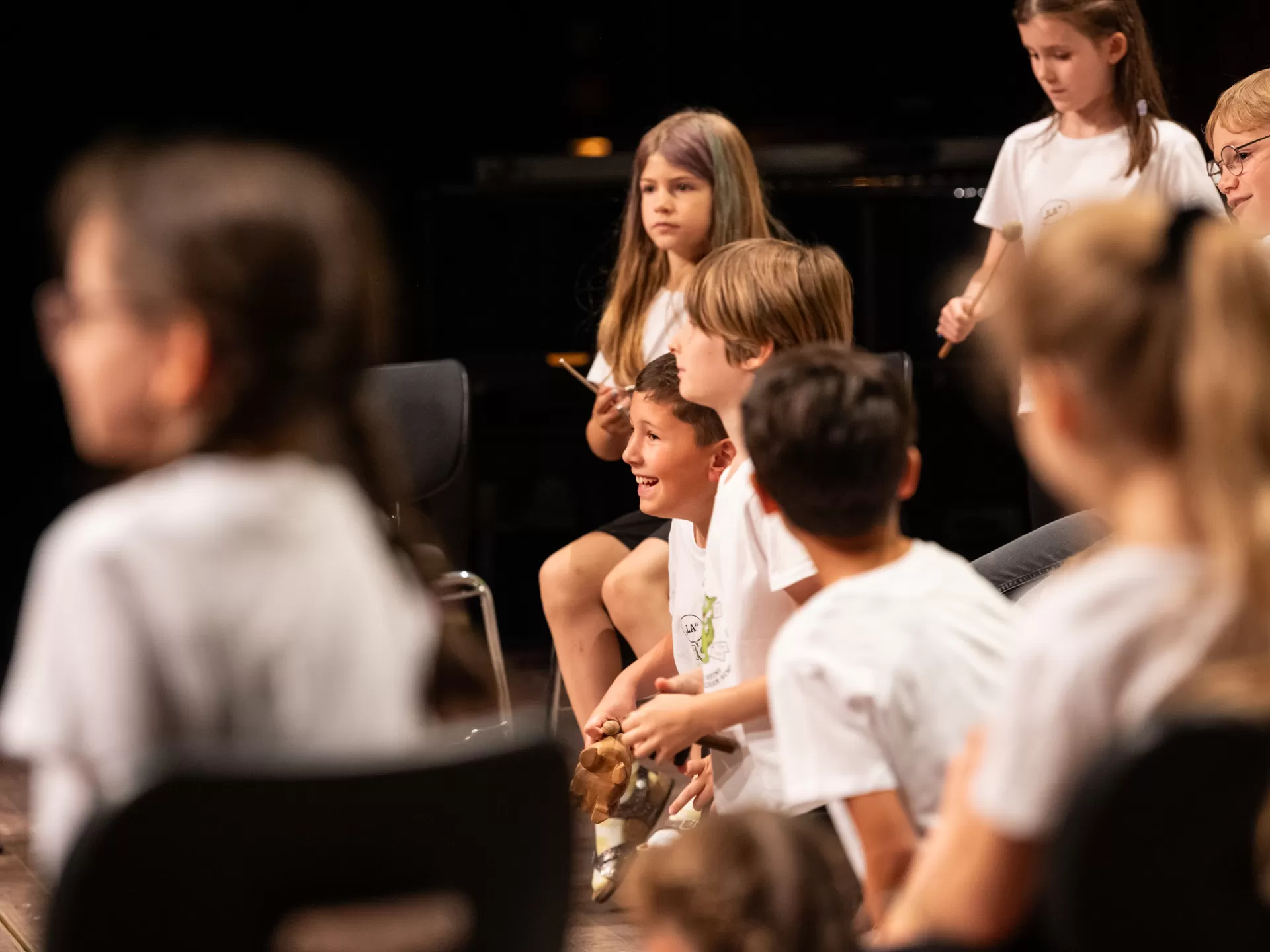 Eine Gruppe Kinder in weißen T-Shirts auf der Bühne. Durch die Kinderschar hindurch fotografiert. Im Fokus steht ein lachender Junge, der halb von einem weiteren Jungen verdeckt wird. Alle blicken nach links.