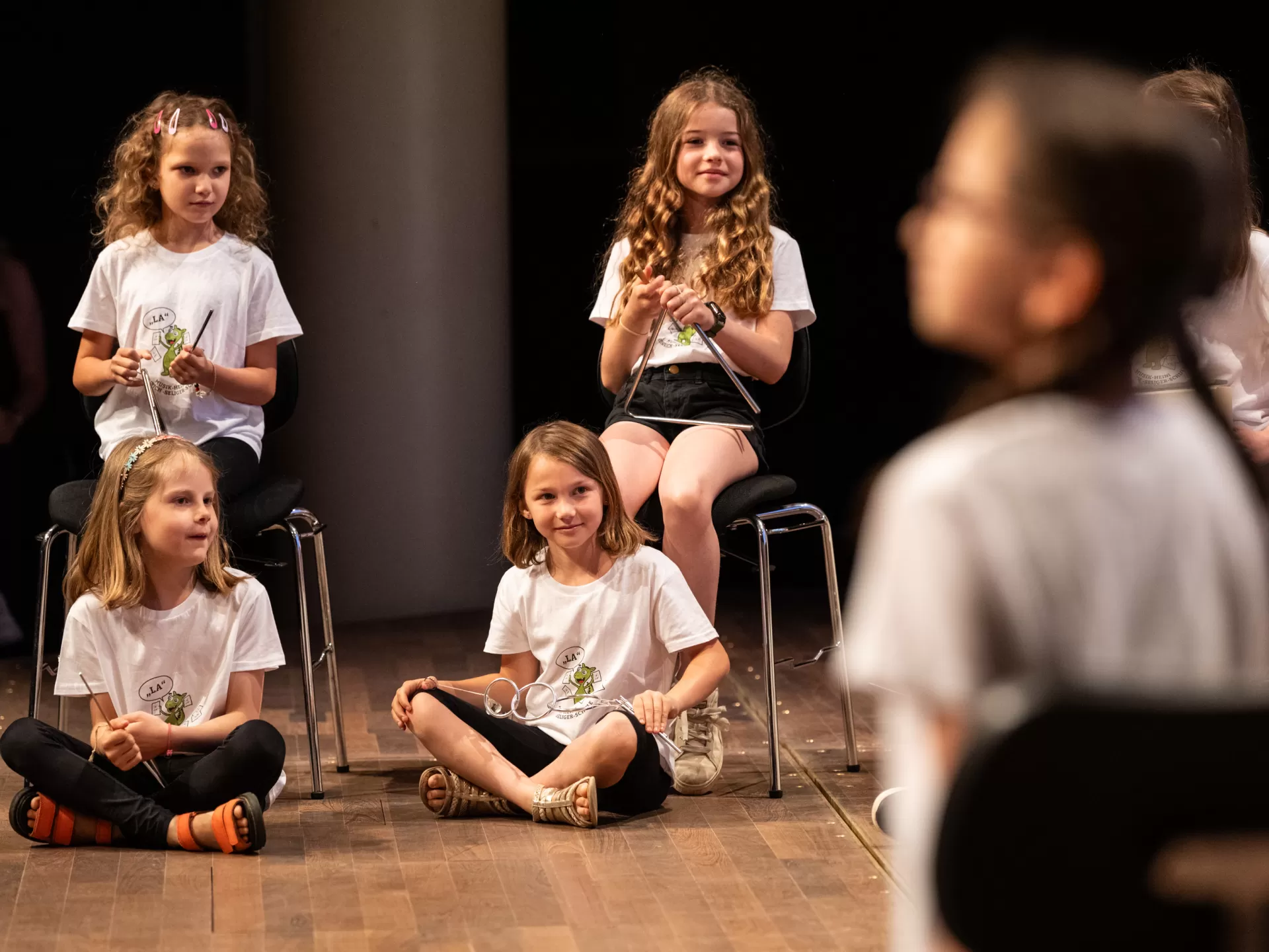 Eine Gruppe Kinder in weißen T-Shirts auf der Bühne. Zu sehen sind hier vier Mädchen. Zwei sitzen auf Stühlen, die anderen beiden davor auf dem Boden. Drei von ihnen halten je eine Triangel. Das vierte Mädchen ein ähnliches Instrument mit drei ineinandergreifenden Ringen. Alle vier blicken lächelnd nach rechts.