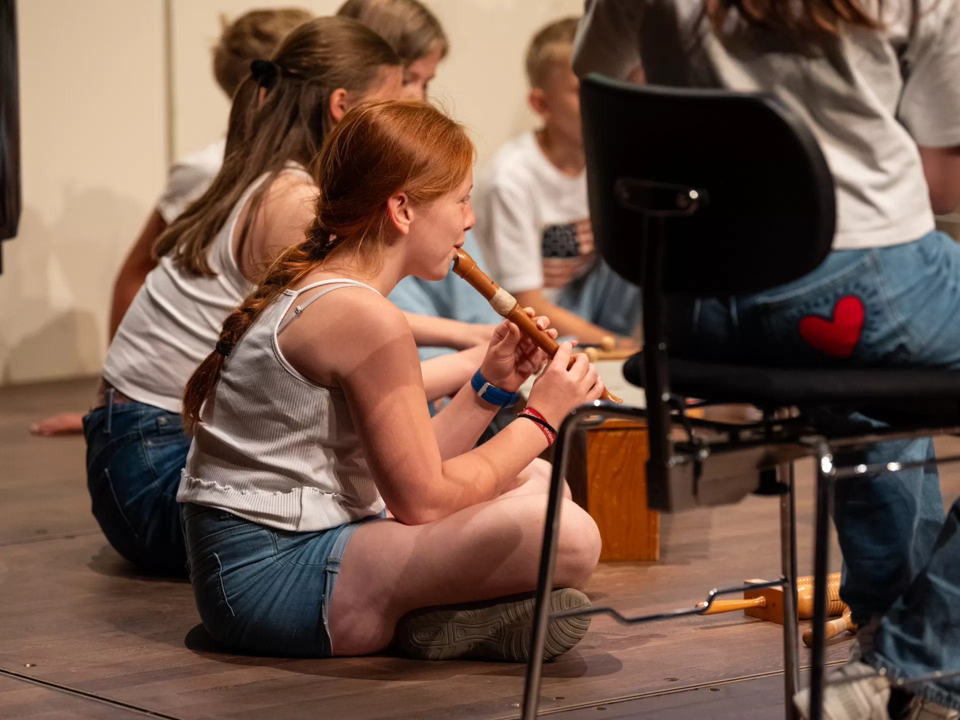 Ein rothaariges Mädchen sitzt im Schneidersitz auf der Bühne und spielt Blockflöte. Sie ist schräg von hinten zu sehen und scheint einem Sitzkreis mit anderen Kindern zu sitzen, von dem nur ein kleiner Teil um sie herum sichtbar ist.