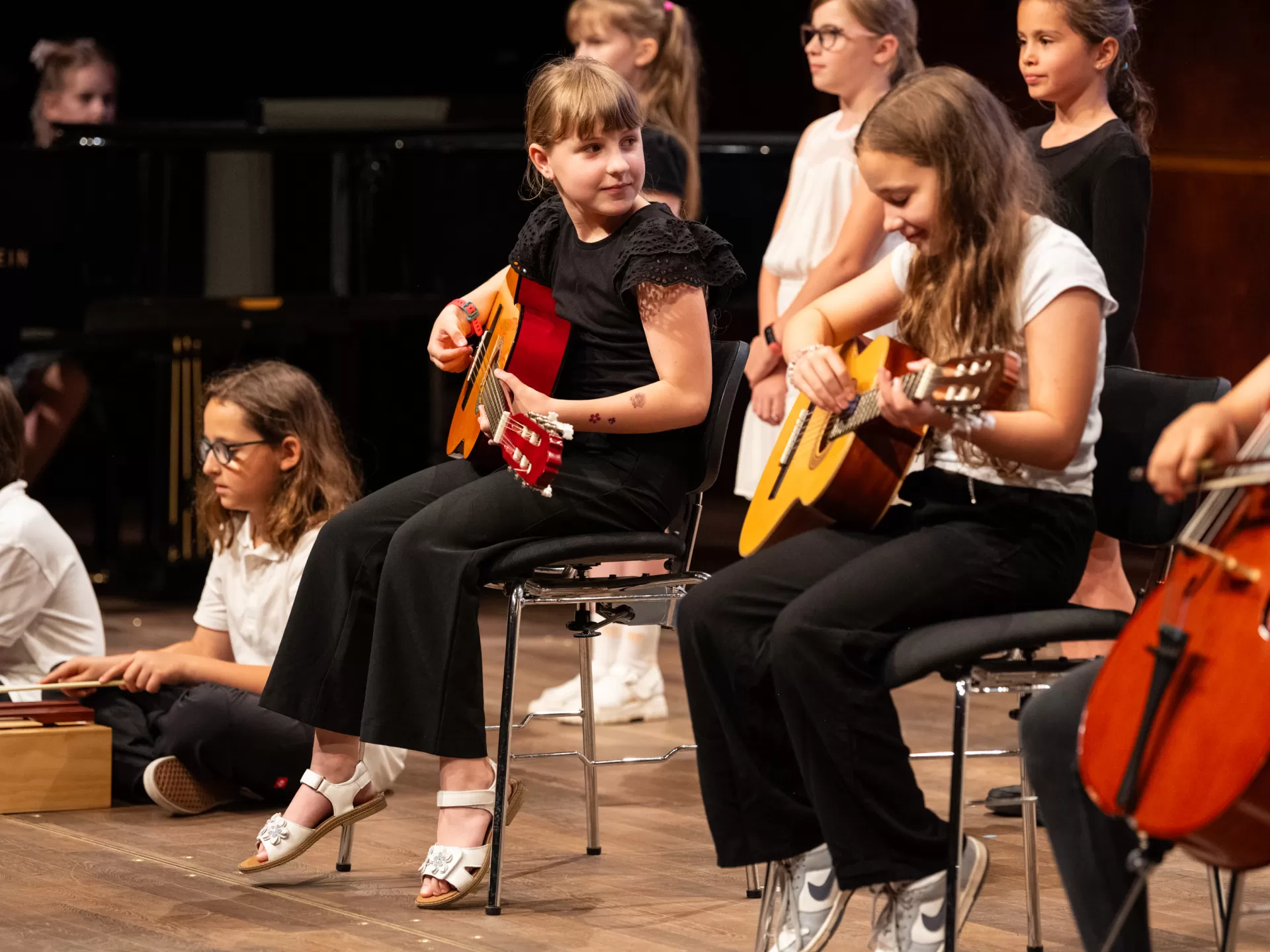 Eine Gruppe Kinder sitzt mit Instrumenten auf der Bühne. Im Fokus stehen zwei Mädchen, die auf Stühlen sitzen und Gitarre spielen. Um sie herum sitzen weitere Kinder, links auf dem Boden eines mit Xylophon, rechts auf einem Stuhl eines mit Cello.