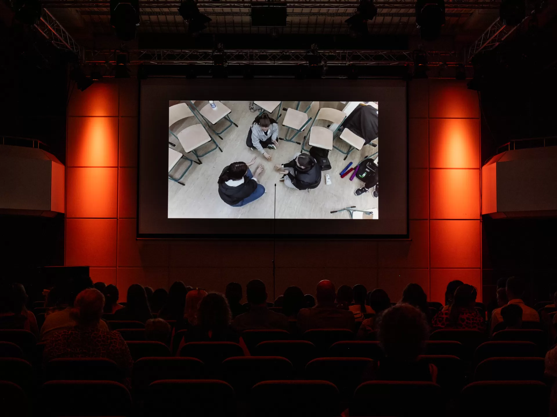 Ein Video wird auf der Leinwand gezeigt. Darin zu sehen sind drei auf dem Boden sitzende Kinder, von oben gefilmt.