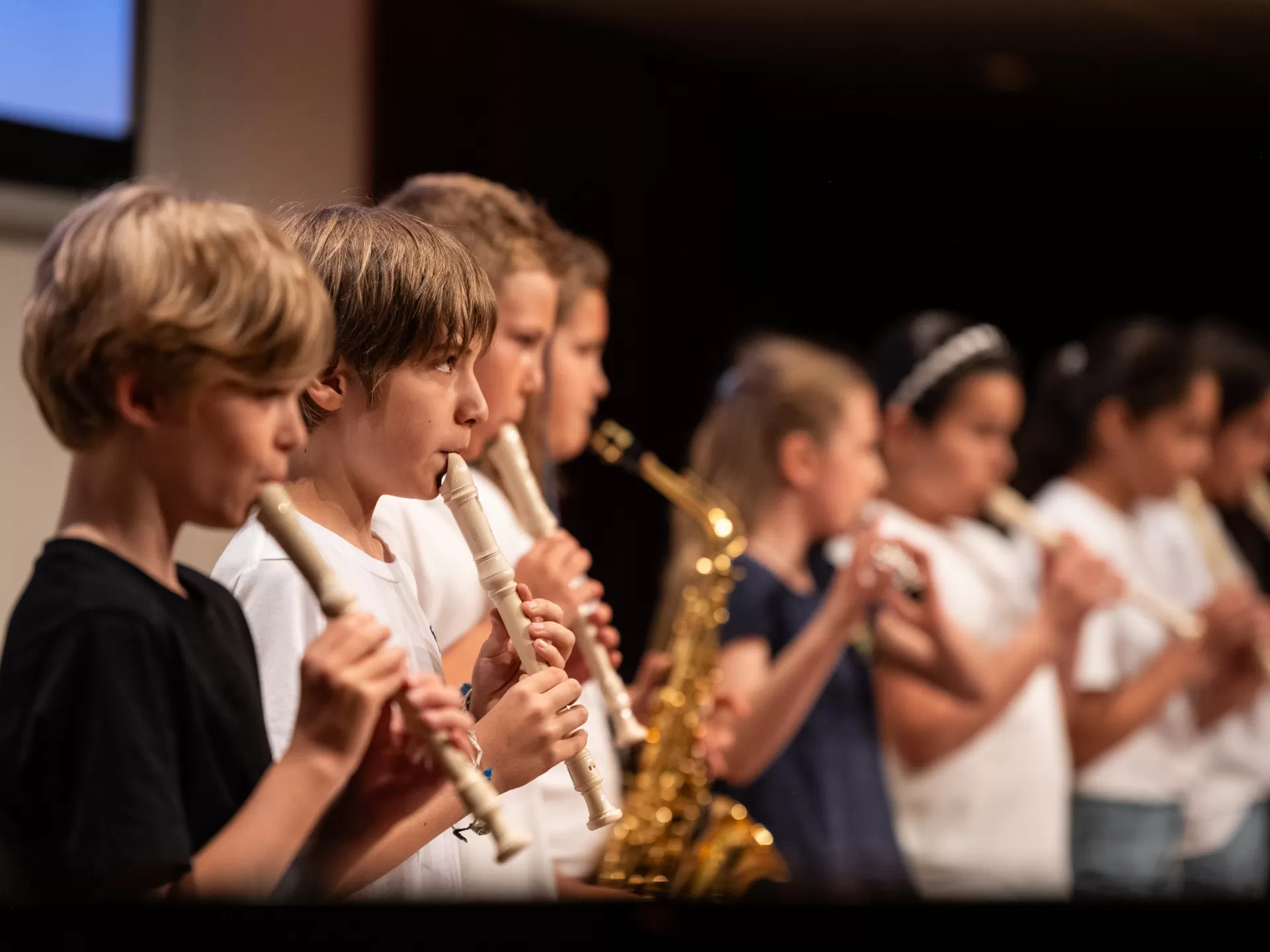 Drei Jungs spielen Blockflöte. Verschwommen im Hintergrund ist zu erkennen, dass weitere Kinder Saxophon und Flöte spielen.