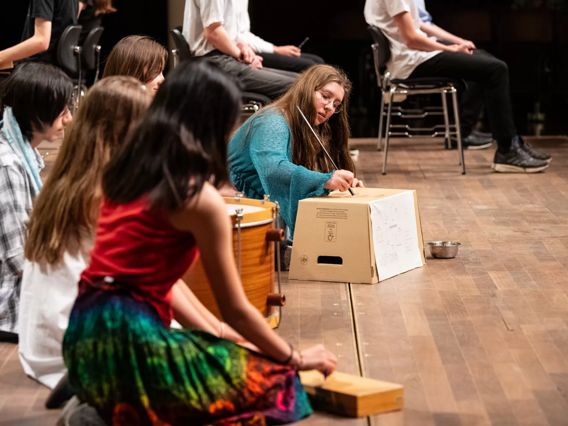 Kinder knien auf dem Boden und spielen auf mehr oder weniger improvisierten Percussion-Instrument. Im Fokus steht ein Mädchen, das mit einem Stock über einen Pappkarton streicht.