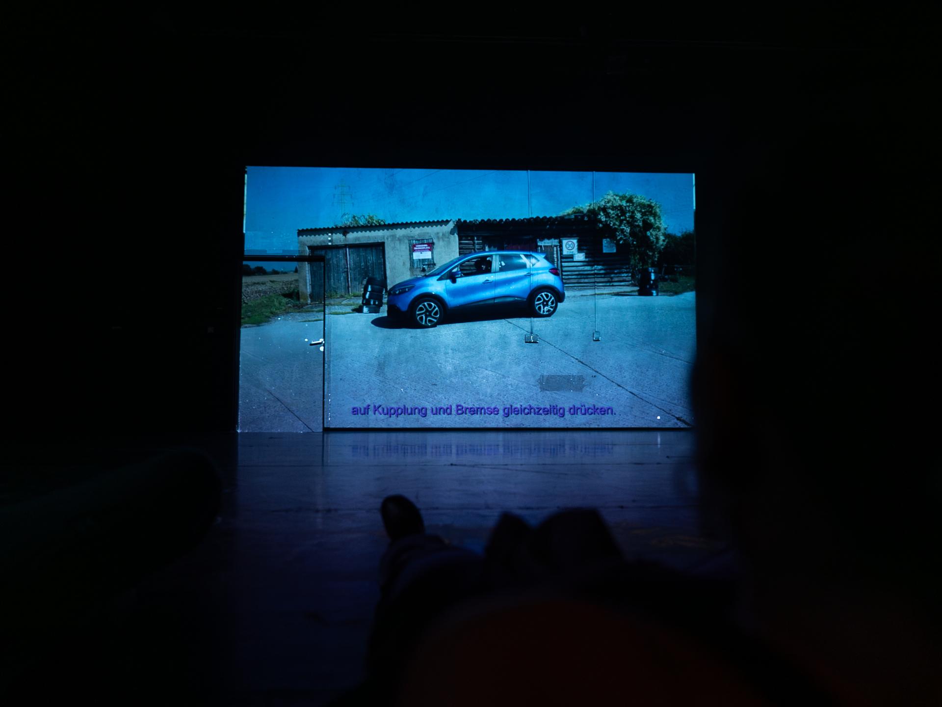 Auf die Rückwand der Bühne ist ein Bild projiziert. Darauf sieht man ein blaues Auto vor einem kleinen Gebäude stehen. Es gibt Untertitel auf den steht: "auf Kupplung und Bremse gleichzeitig drücken."