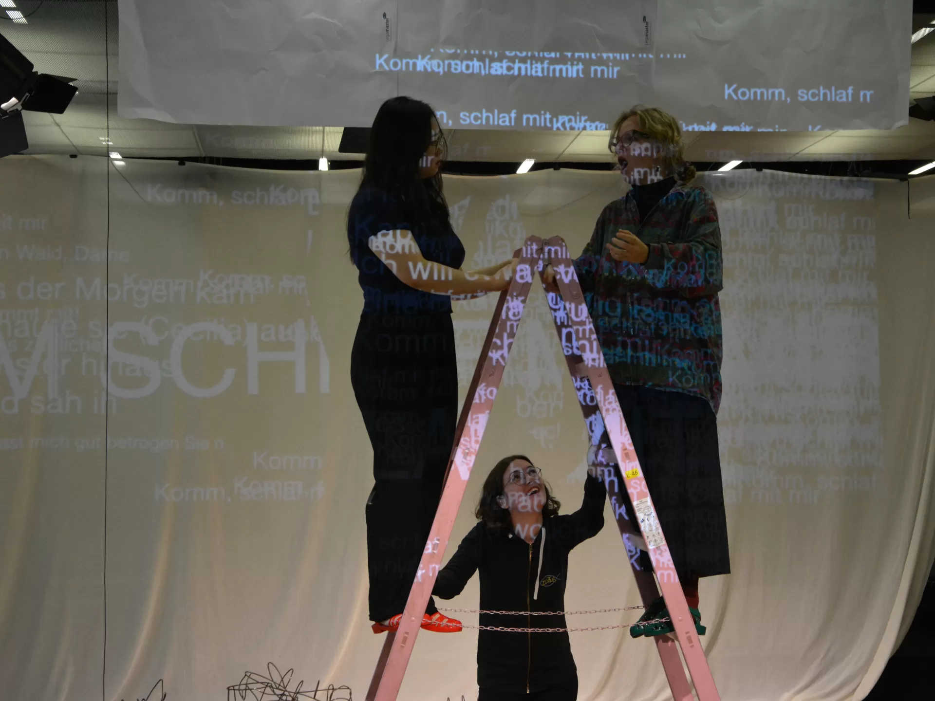 Probenfoto: Zwei Sängerinnen stehen auf einer Leiter. Eine dritte steht unten und schaut zu ihnen hoch.