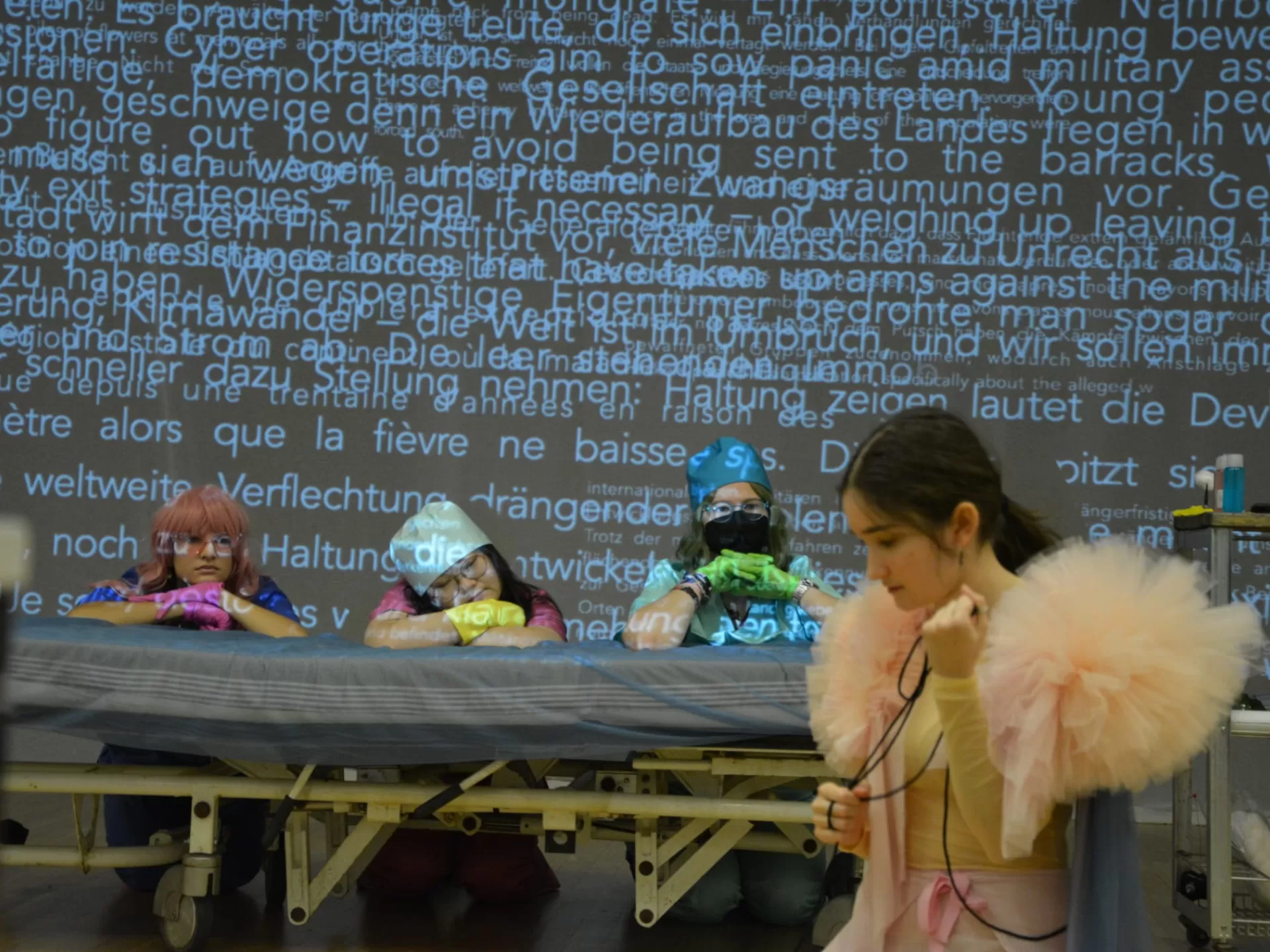 Probenfoto für "Schweig still, mein Stein": Im Vordergrund kniet die Tänzerin und blickt auf den Boden. Die drei Sängerinnen knien hinter einem Krankenhausbett und schauen sie an. Dahinter befindet sich eine Leinwand, auf die eine große Menge Text projiziert ist.
