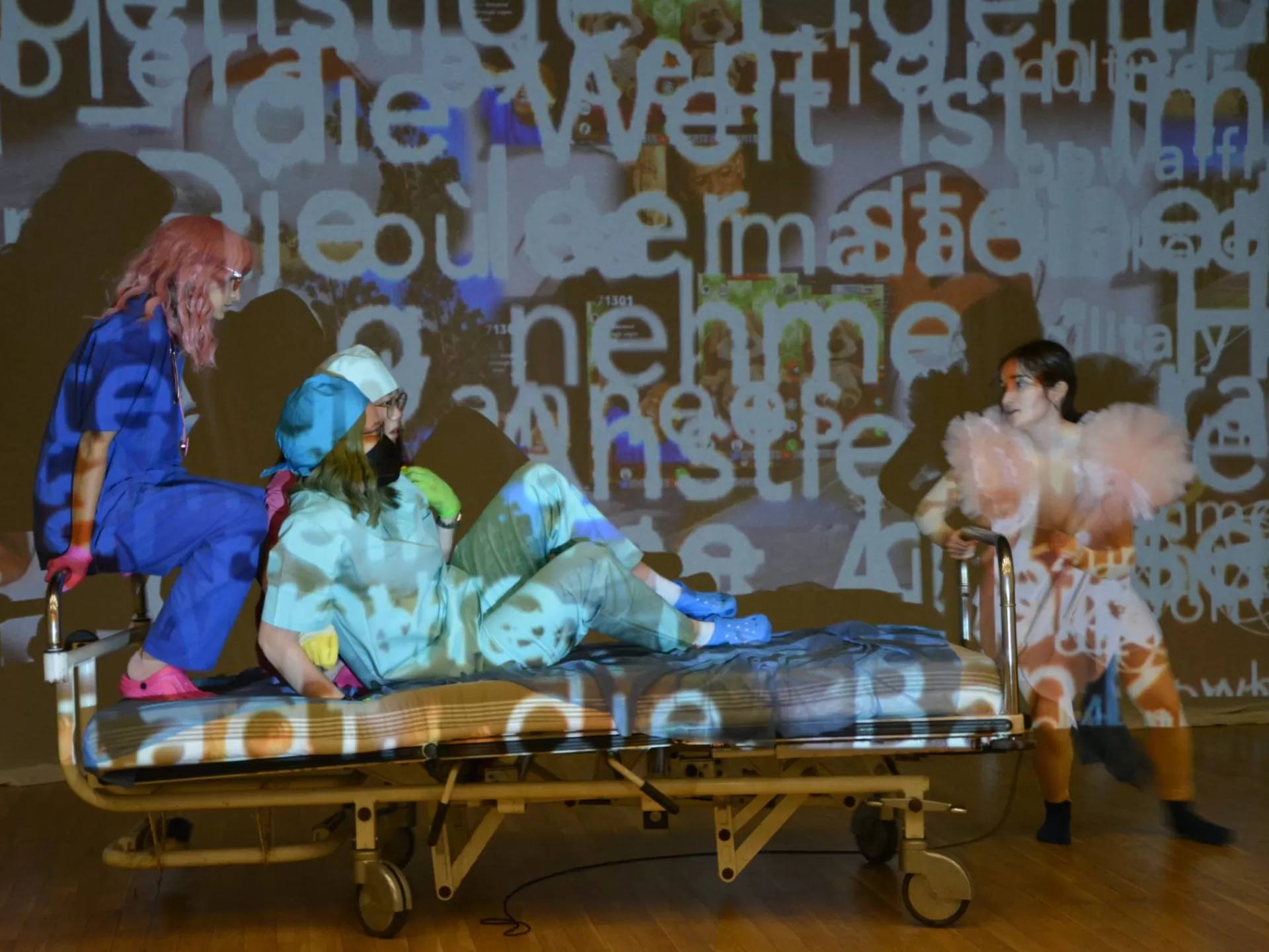 Probenfoto: Drei Darstellerinnen sitzen im Krankenbett, welches von einer vierten gezogen wird. Sie und die Leinwand im Hintergrund werden von einer Projektion mit Text angeleuchtet.