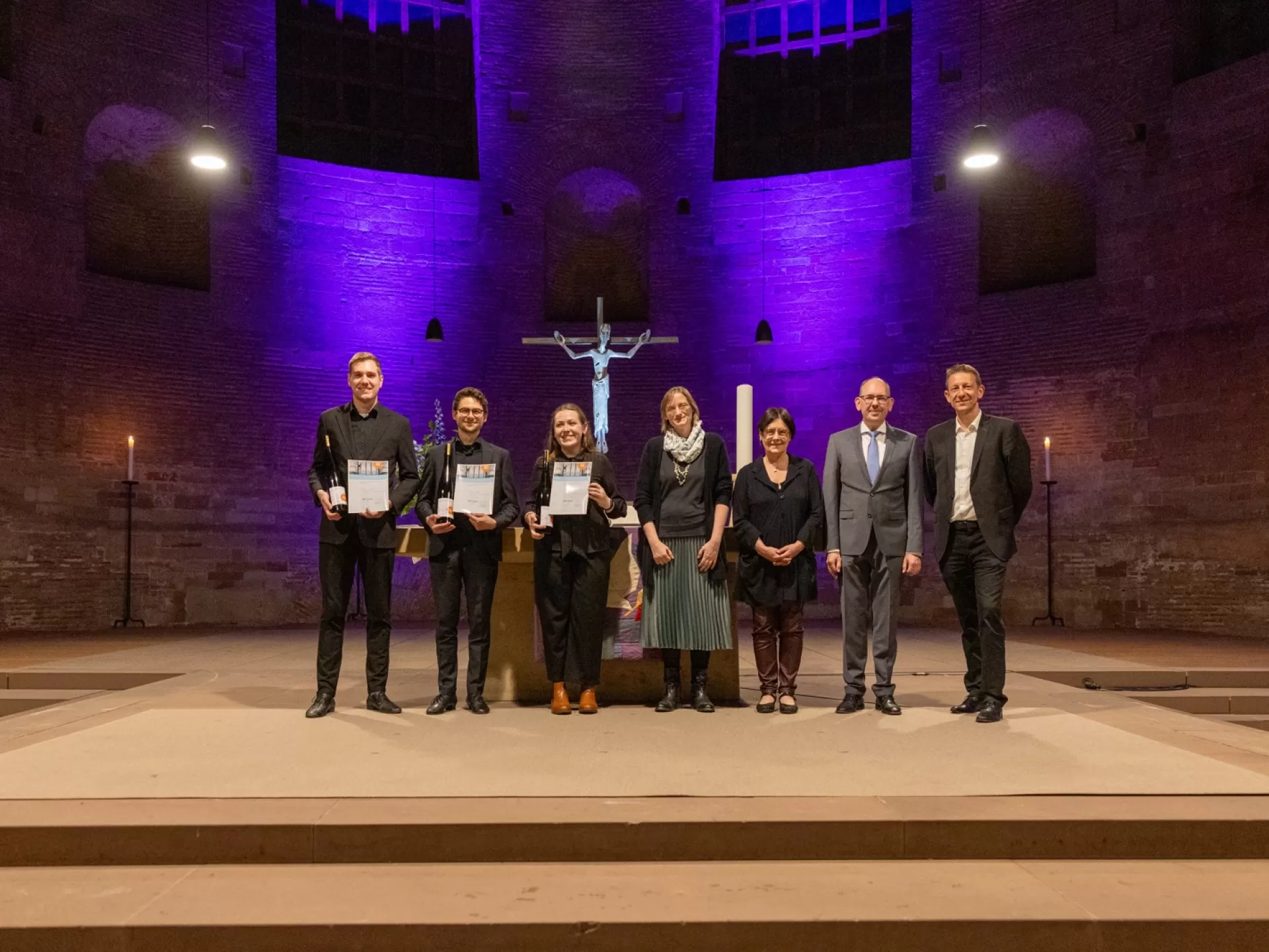 Sieben Personen in formeller Kleidung stehen in einer Reihe in einer Kirche. Die drei Preisträger*innen stehen links und präsentieren ihre Urkunden.