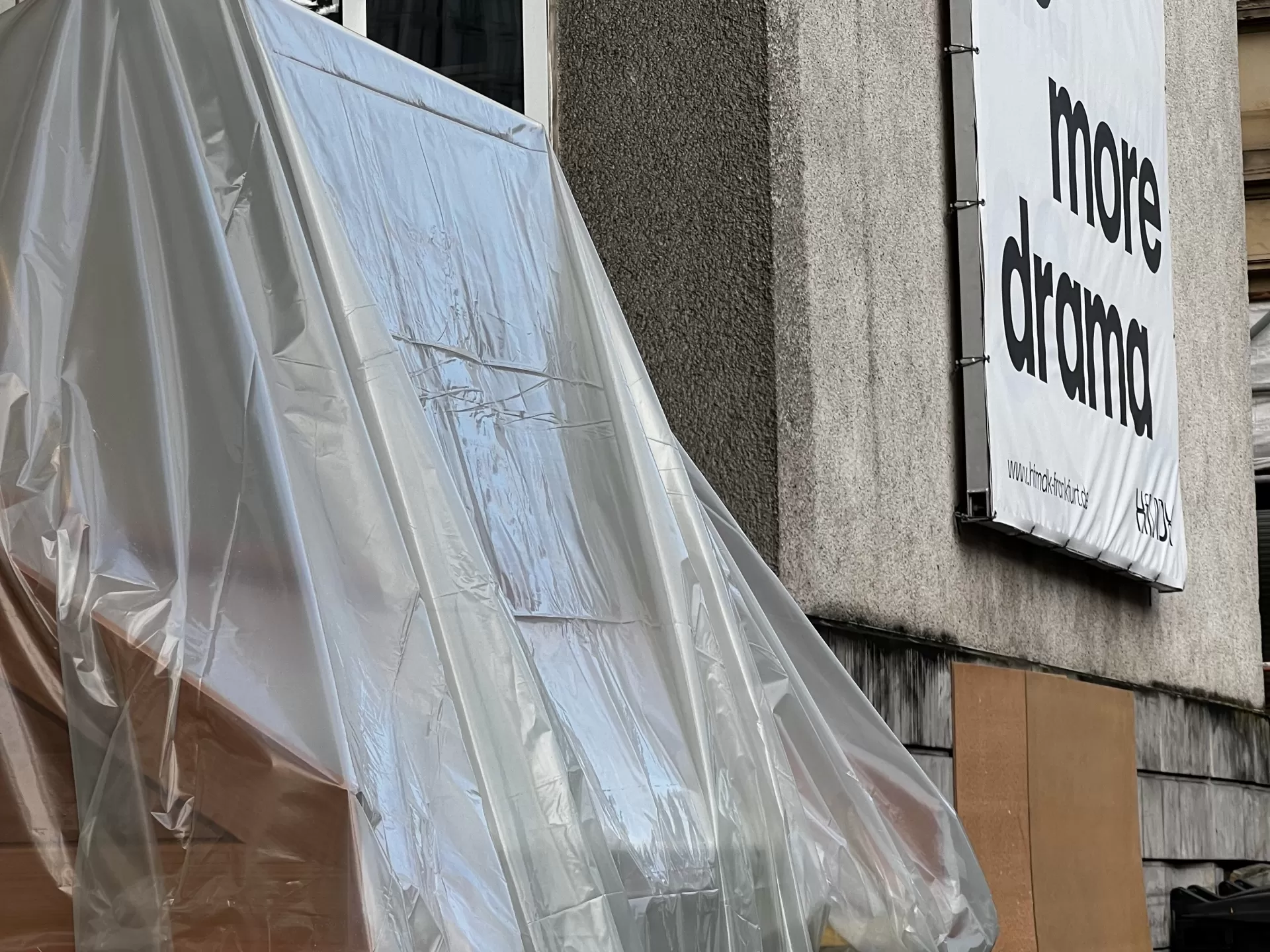 Der unter einer Plane versteckte Spieltisch steht vor dem Hauptgebäude der HfMDK. Im Hintergrund sieht man das Fassadenbanner mit der Aufschrift: "no more drama", dabei ist das Wort "no" durchgestrichen.