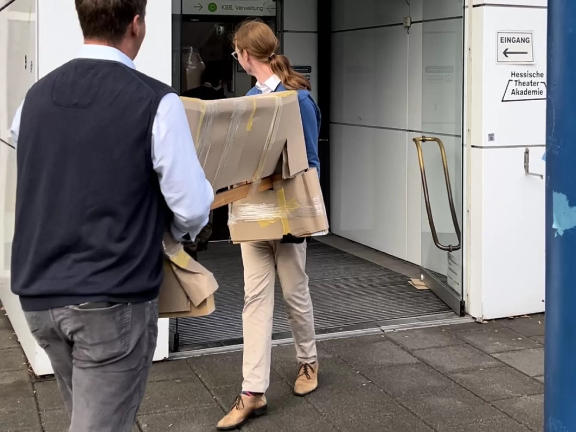 Zwei Personen tragen ein längliches Paket durch den Haupteingang der Hochschule.