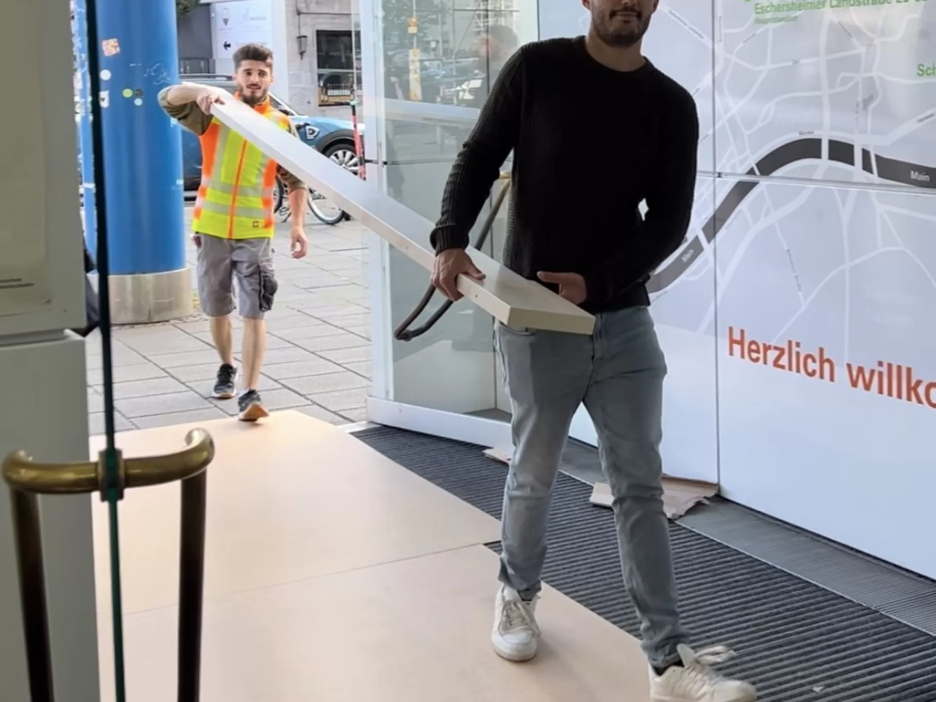 Zwei Personen tragen ein langes Brett durch den Haupteingang.