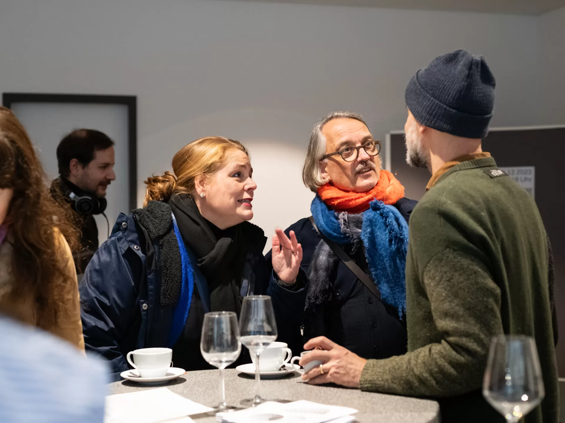 Mehrere Personen in einem Foyer, drei Personen unterhalten sich an einem Stehtisch bei Kaffee und Wasser