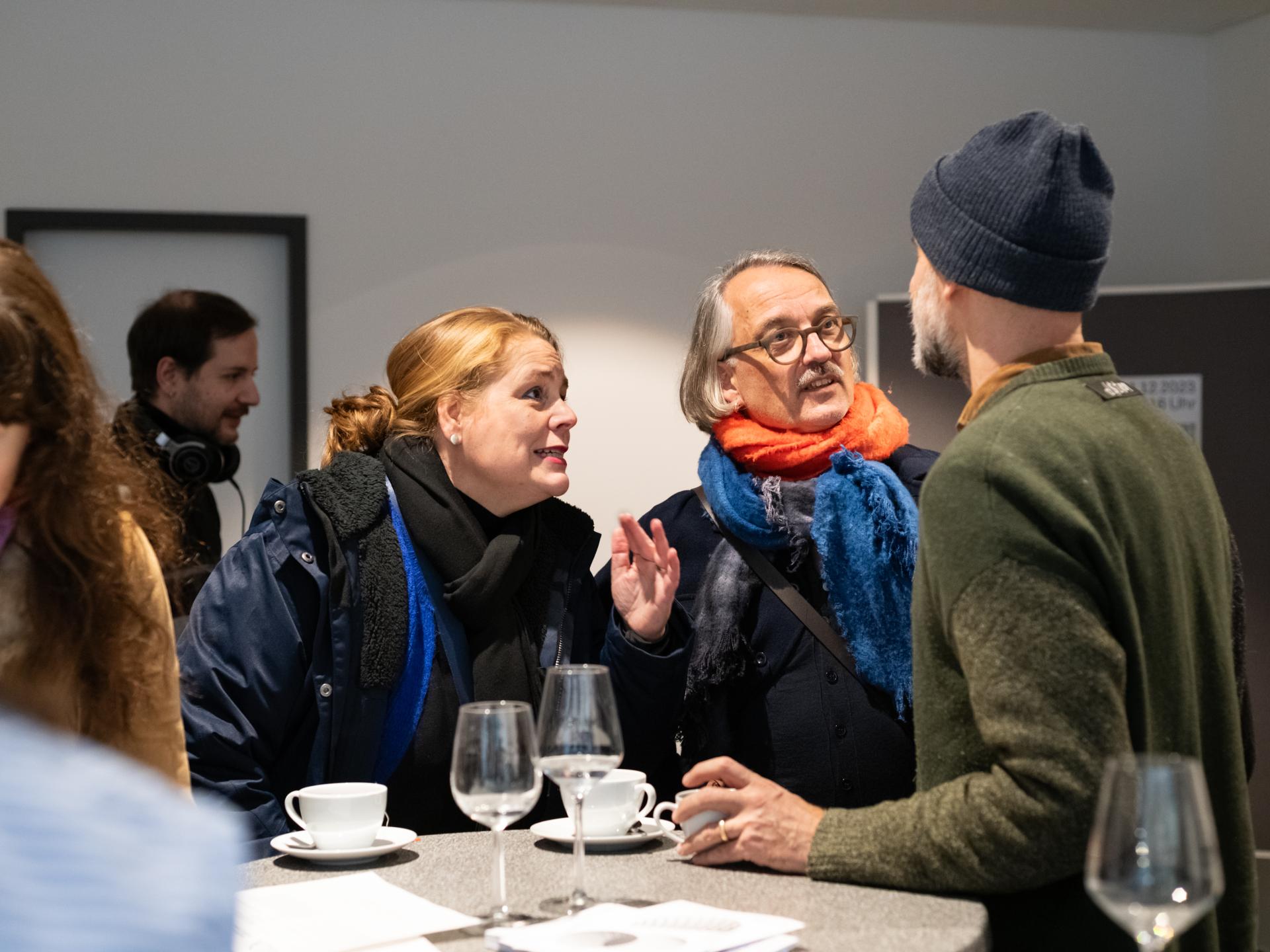 Mehrere Personen in einem Foyer, drei Personen unterhalten sich an einem Stehtisch bei Kaffee und Wasser