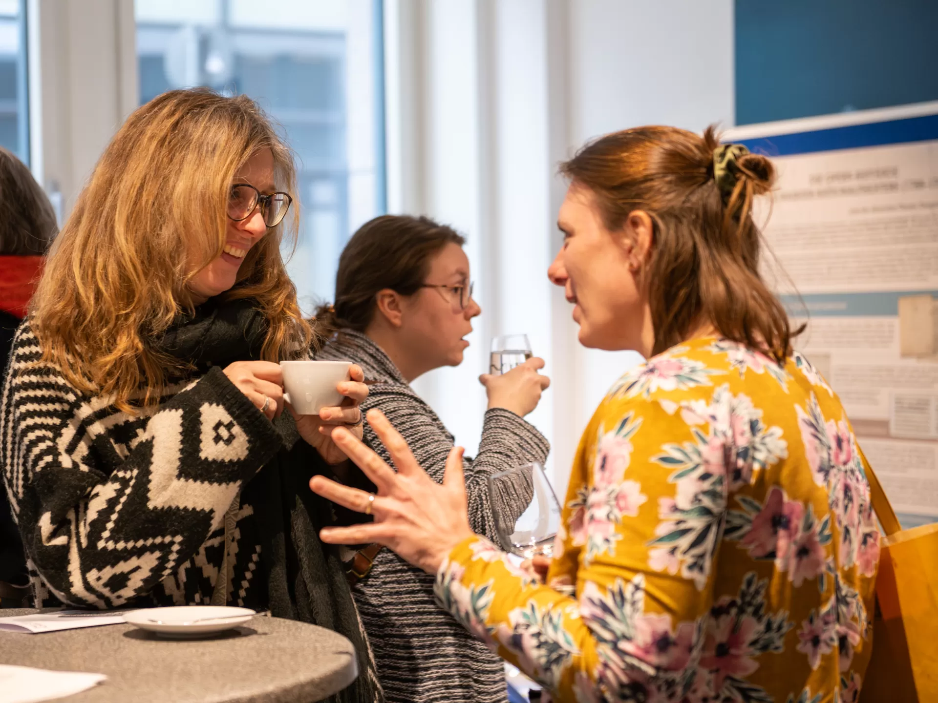 Zwei Personen unterhalten sich an einem Stehtisch bei einer Tasse Kaffee, im Hintergrund weitere Personen und Schautafeln