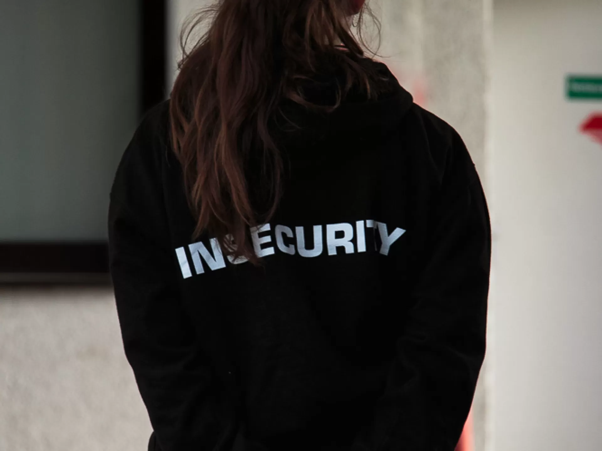 Eine Person mit langem Pferdeschwanz ist stehend von hinten zu sehen, bis Hüfthöhe, sie hält die Hände hinter dem Körper zusammen. Sie ist komplett schwarz gekleidet, auf ihrem schwarzen Hoodie steht in weiß "insecurity". 
