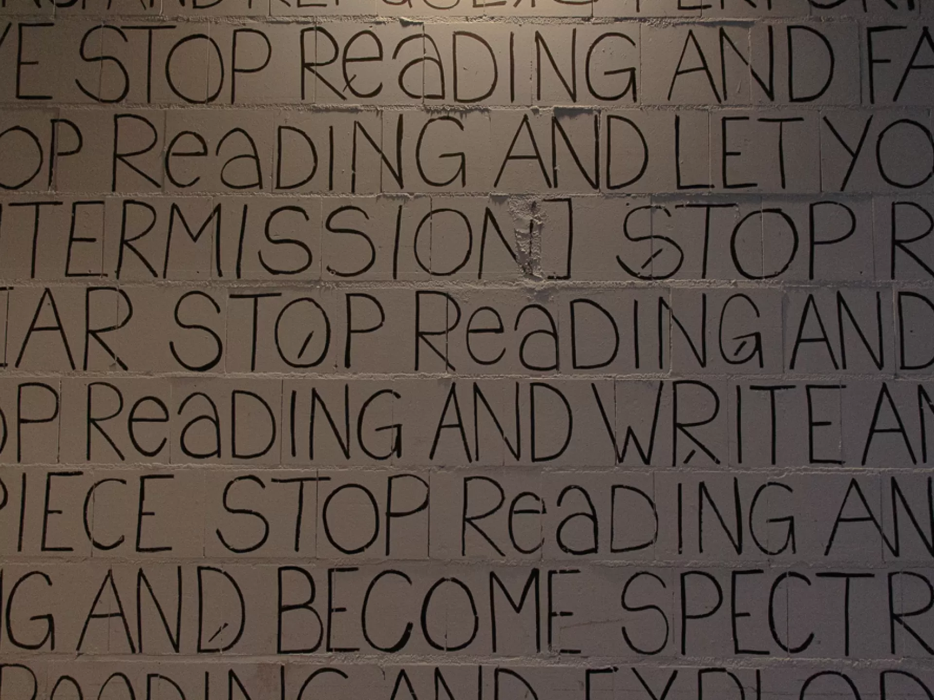 Auf einer weißen Mauerwand stehen in schwarzer Schrift englische Worte, es sind mehrere dicht geschriebene Zeilen zu sehen. In dem Ausschnitt kann man lesen "intermission stop", "stop reading", "stop readking and write", "and become spectra..."