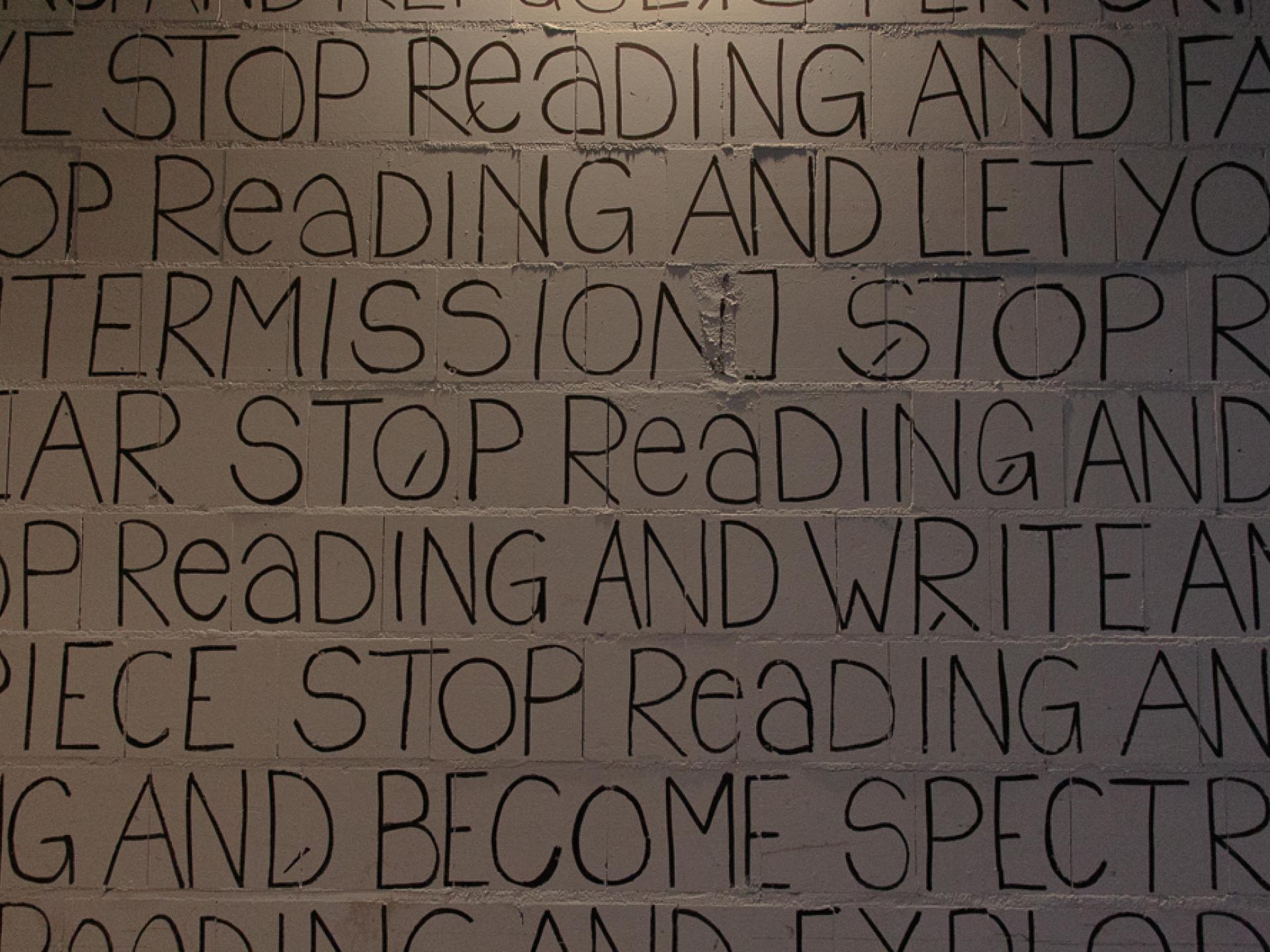Auf einer weißen Mauerwand stehen in schwarzer Schrift englische Worte, es sind mehrere dicht geschriebene Zeilen zu sehen. In dem Ausschnitt kann man lesen "intermission stop", "stop reading", "stop readking and write", "and become spectra..."