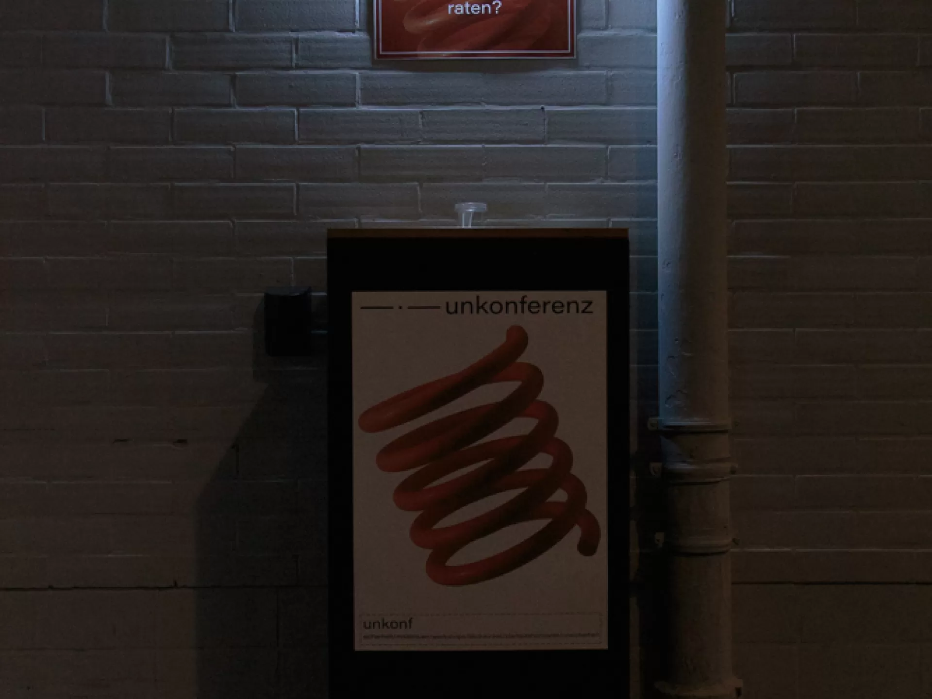 Vor einer weißen Mauerwand steht ein Aufsteller mit einem Poster auf dem der Titel "unkonferenz" zu lesen ist und ein spiralförmiges Logo. Auf dem Boden davor sind drei Fragezeichen zu sehen. An der Wand hängt ein kleines Poster, das angeleuchtet wird, darauf steht "was würdest du deinem jüngeren ich raten?".