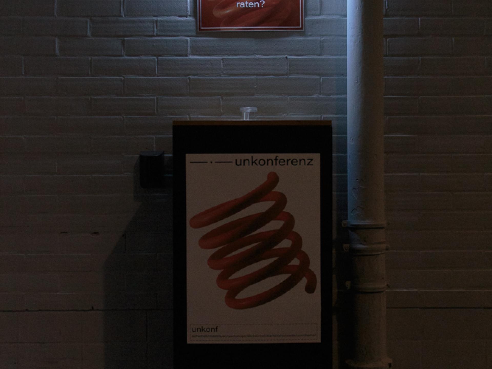 Vor einer weißen Mauerwand steht ein Aufsteller mit einem Poster auf dem der Titel "unkonferenz" zu lesen ist und ein spiralförmiges Logo. Auf dem Boden davor sind drei Fragezeichen zu sehen. An der Wand hängt ein kleines Poster, das angeleuchtet wird, darauf steht "was würdest du deinem jüngeren ich raten?".