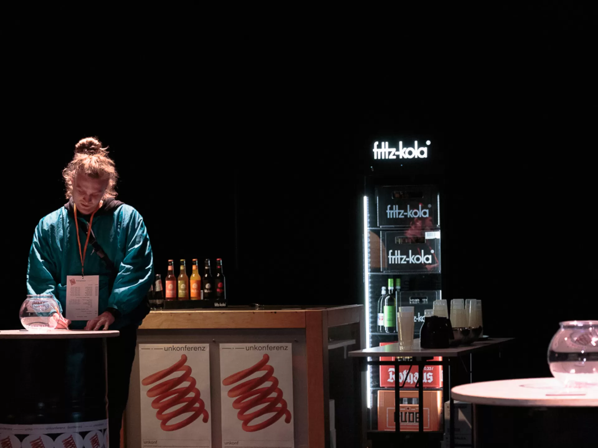 Eine Frau steht hinter einem Tisch, sie ist bis Hüfthöhe zu sehen und träge eine blaue Jacke und trägt an einem Band um den Hals einen Zettel. Vorn am Tisch ist ein Poster mit dem Titel "unkonferenz" und einem spiralförmigen Loge. Neben ihr auf dem Tisch stehen kleine Getränkeflaschen mit Limonade. Rechts neben dem Tisch steht ein weiterer, niedrigerer Tisch mit Getränkebecher, dahinter ein Glaskühlschrank von "fritz-kola" mit Flaschen darin. Rechts davon ein runter Tisch mit einer runden Glasvase darauf.