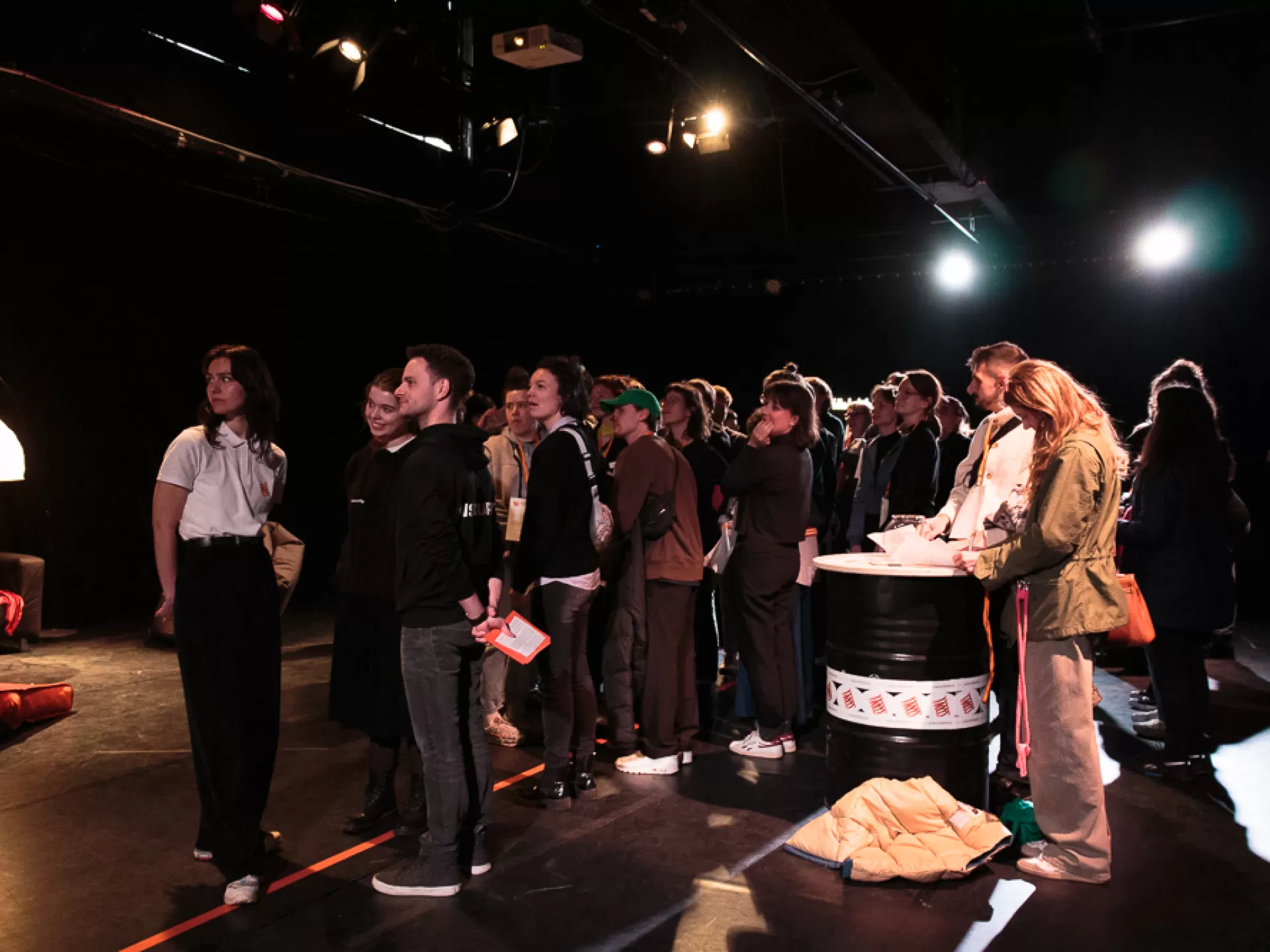 Rechts im Bild steht eine runde Tonne wie ein Empfangstisch mit zwei Leuten dahinter. Links davon steht eine kleine Menschenmenge, alle blicken nach links aus dem Bild, sie beobachten etwas und scheinen zu warten.