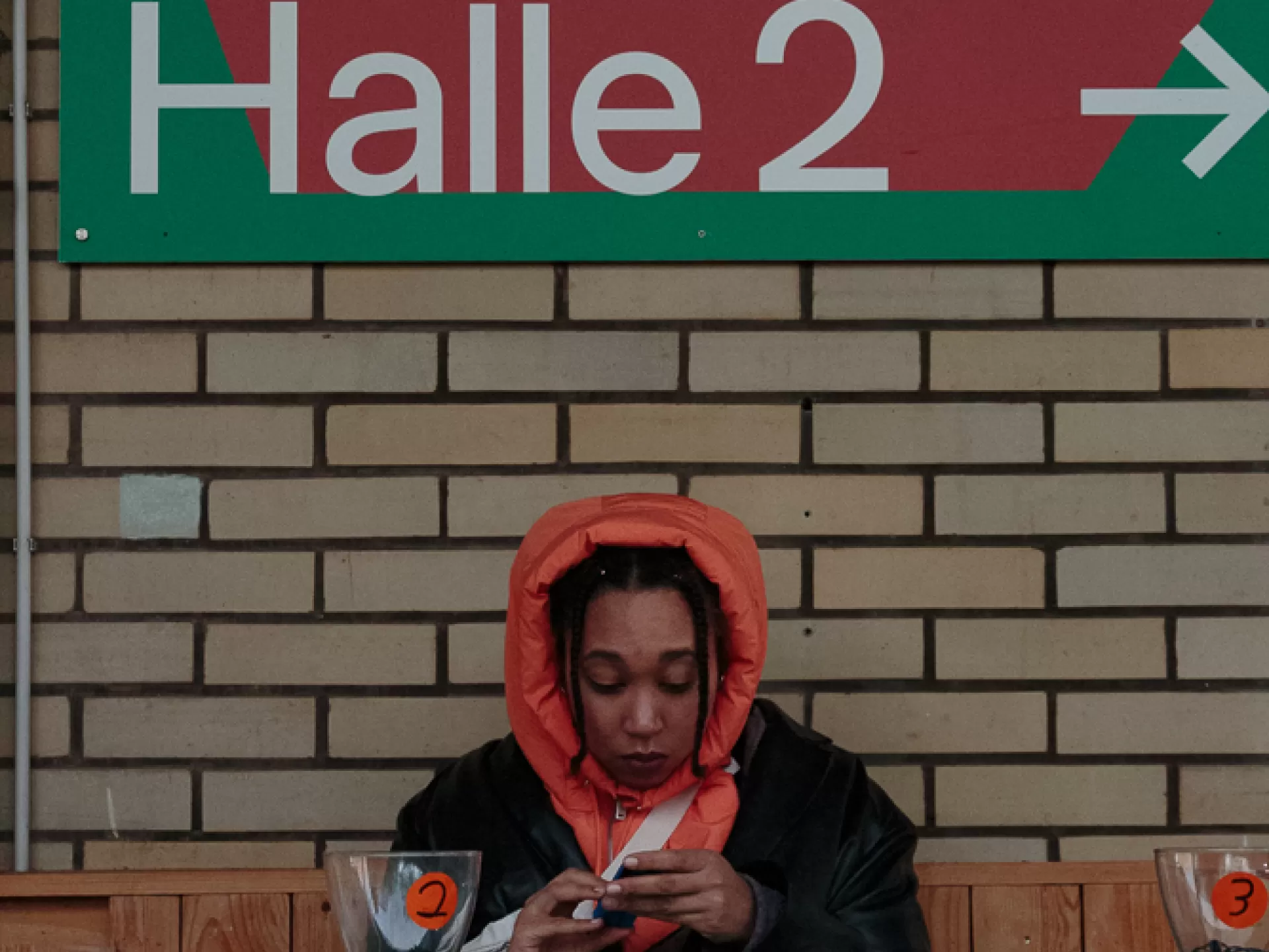 Eine Person sitzt an einem Tisch, hält einen kleinen Gegenstand in den Händen und betrachtet ihn. Auf dem Tisch vor ihr liegt eine Baseballkappe und ein weißer Kopfhörer. Links und ganz rechts auf dem Tisch steht jeweils ein Glasgefäß, auf dem linken ist die Nummer 2, auf dem rechten die Nummer 3 zu sehen.