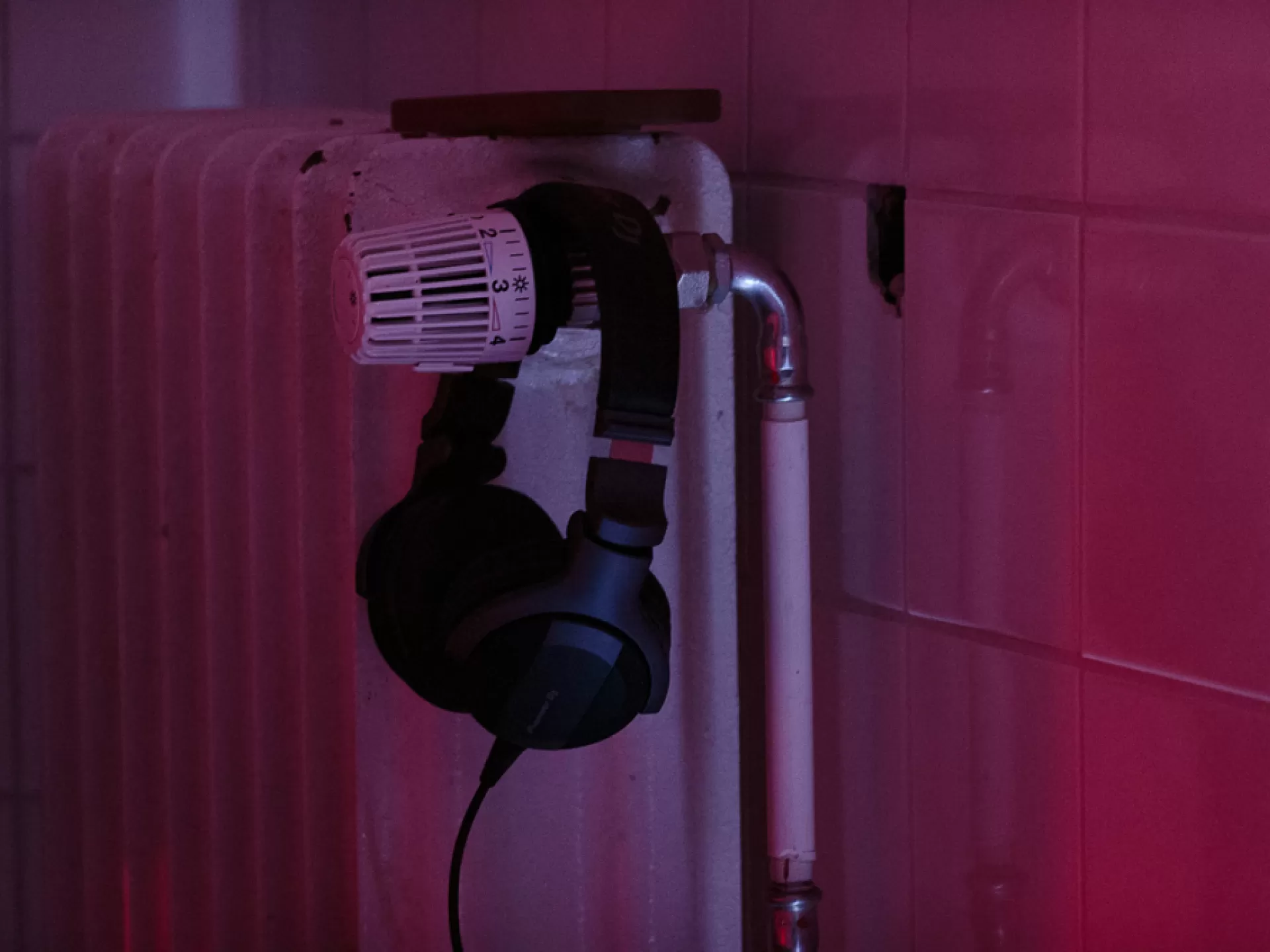 Ein Kopfhöhrer hängt über dem Thermostat eines Heizkörpers vor einer Kachelwand die Szene ist in rosa Licht getaucht.