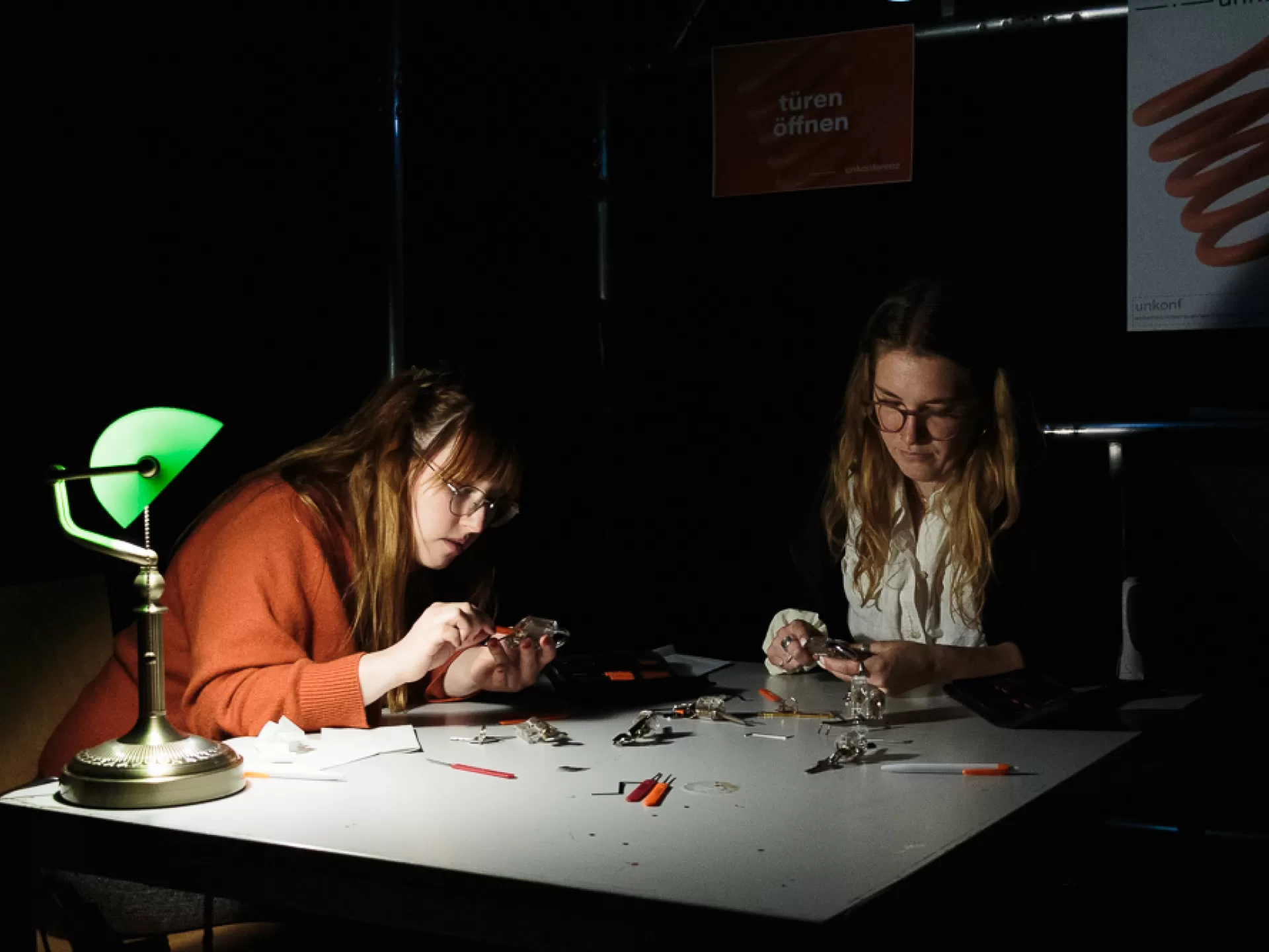 Zwei Frauen sitzen an einem Tisch und arbeiten mit den Händen an kleinen Metallgegenständen, links auf dem Tisch steht eine grüne Bibliothekslampe, auf dem Tisch liegt ein Kuli und kleine Arbeitswerkzeuge.
