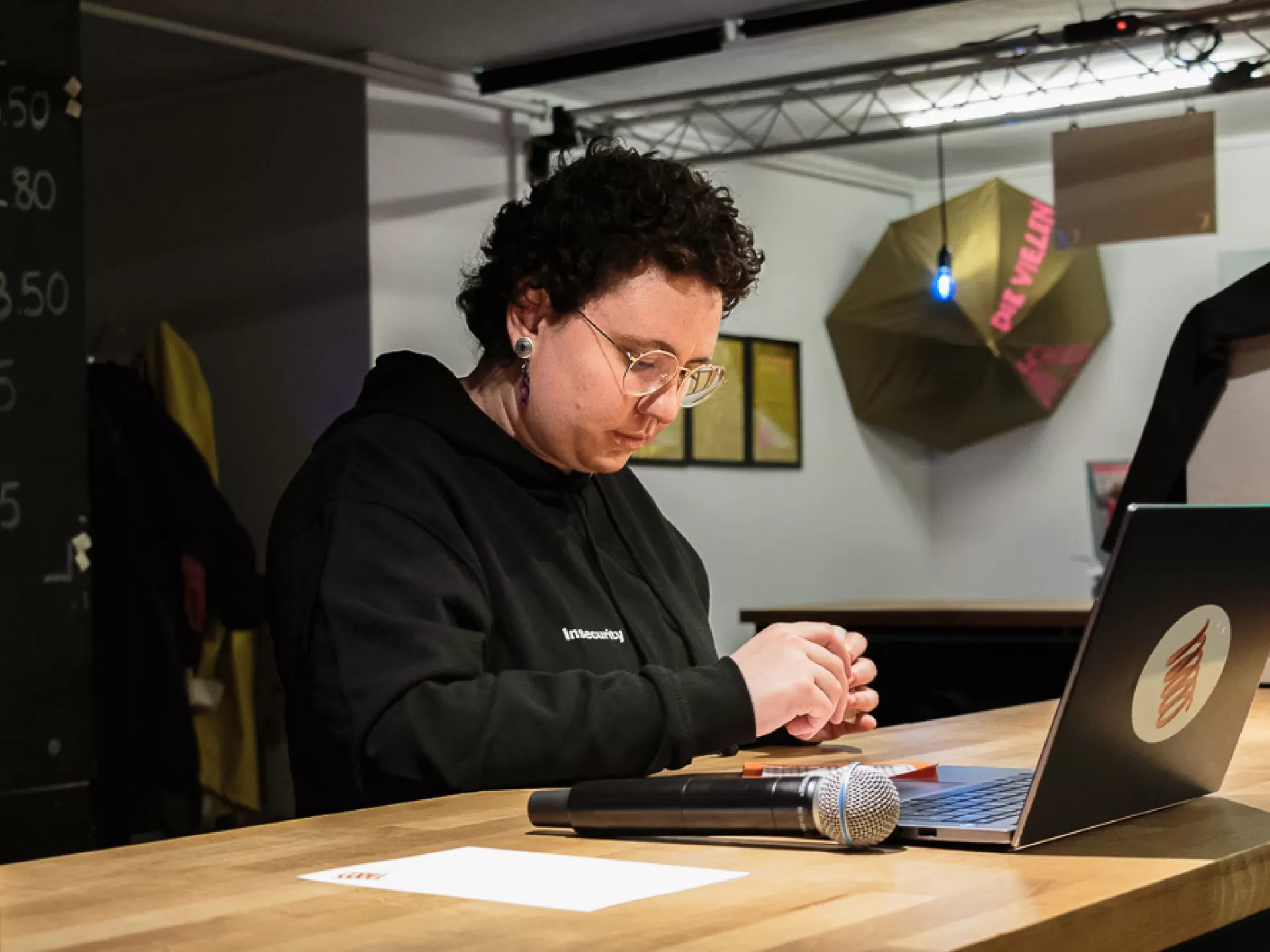 Eine Frau sitzt an einem Tisch, sie trägt einen schwarzen Hoodie und hält die Hände vor ihrem Oberkörper über dem Tisch und blickt nachdenklich nach unten. Auf dem Tisch rechts vor ihr befindet sich ein Laptop, ein Mikrofon und ein weißes Blatt Papier.