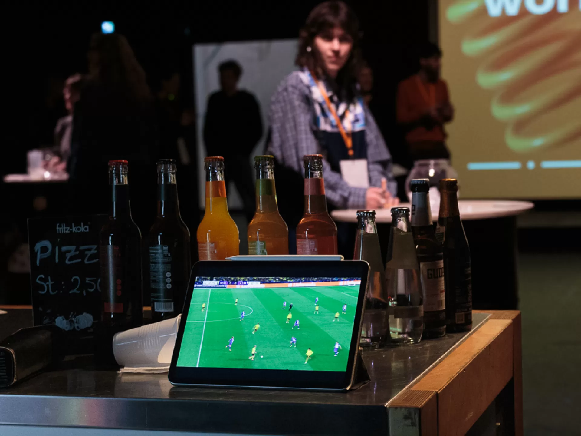 Im Vordergrund steht ein iPad auf dem ein Fußballspiel zu sehen ist, dahinter mehrere kleine Glasflaschen mit Getränken wie Limonade und Wasser. Auf einem kleinen Schild steht "Pizza St. 2,50". Im Hintergrund sind im Raum unscharf Menschen zu sehen.